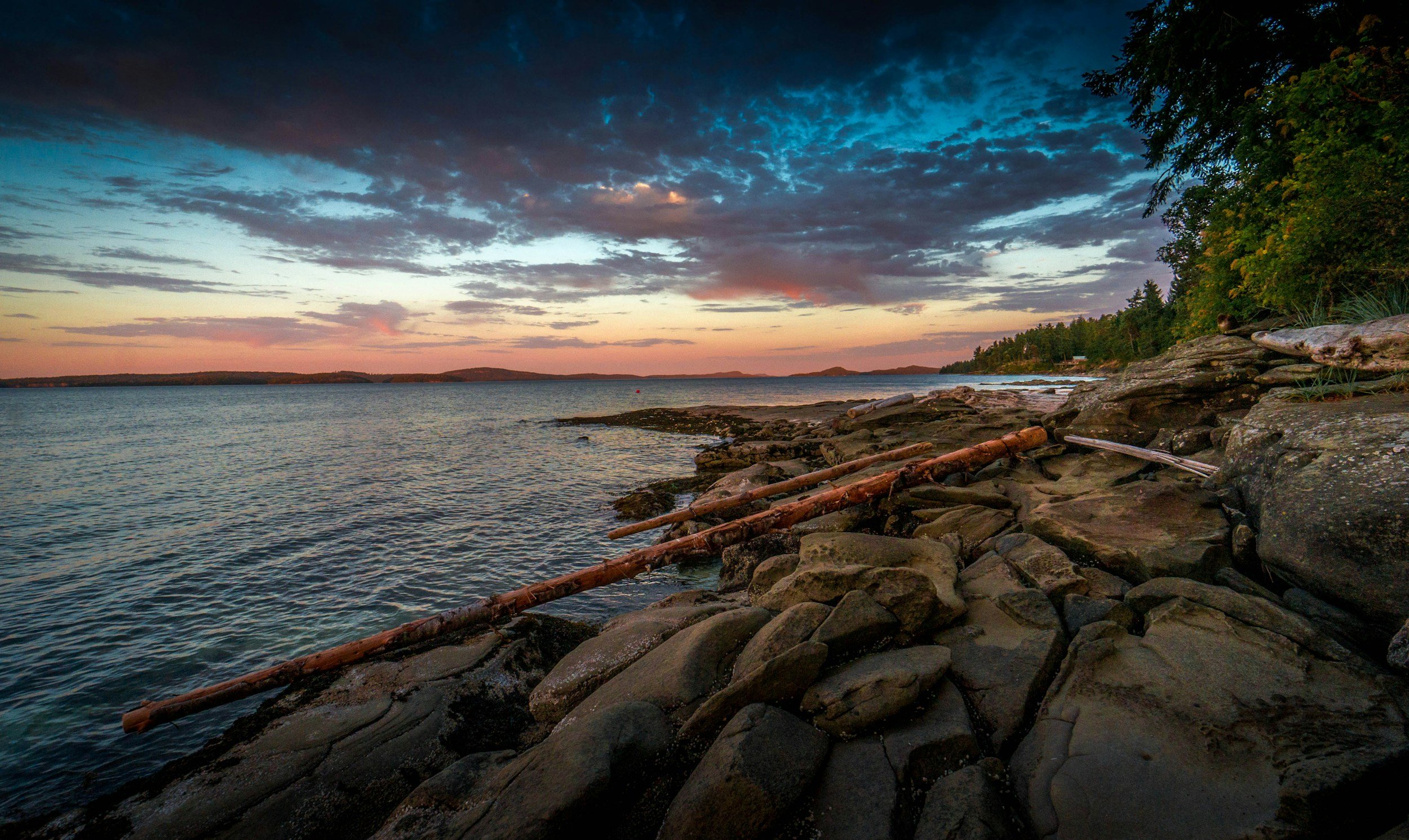 A lakeshore at sunset with a colorful sky, rocky shoreline, driftwood, and trees.