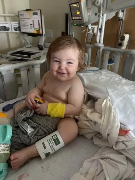 A smiling young child with a wristband, sitting in a hospital bed, holding a toy, surrounded by medical equipment.