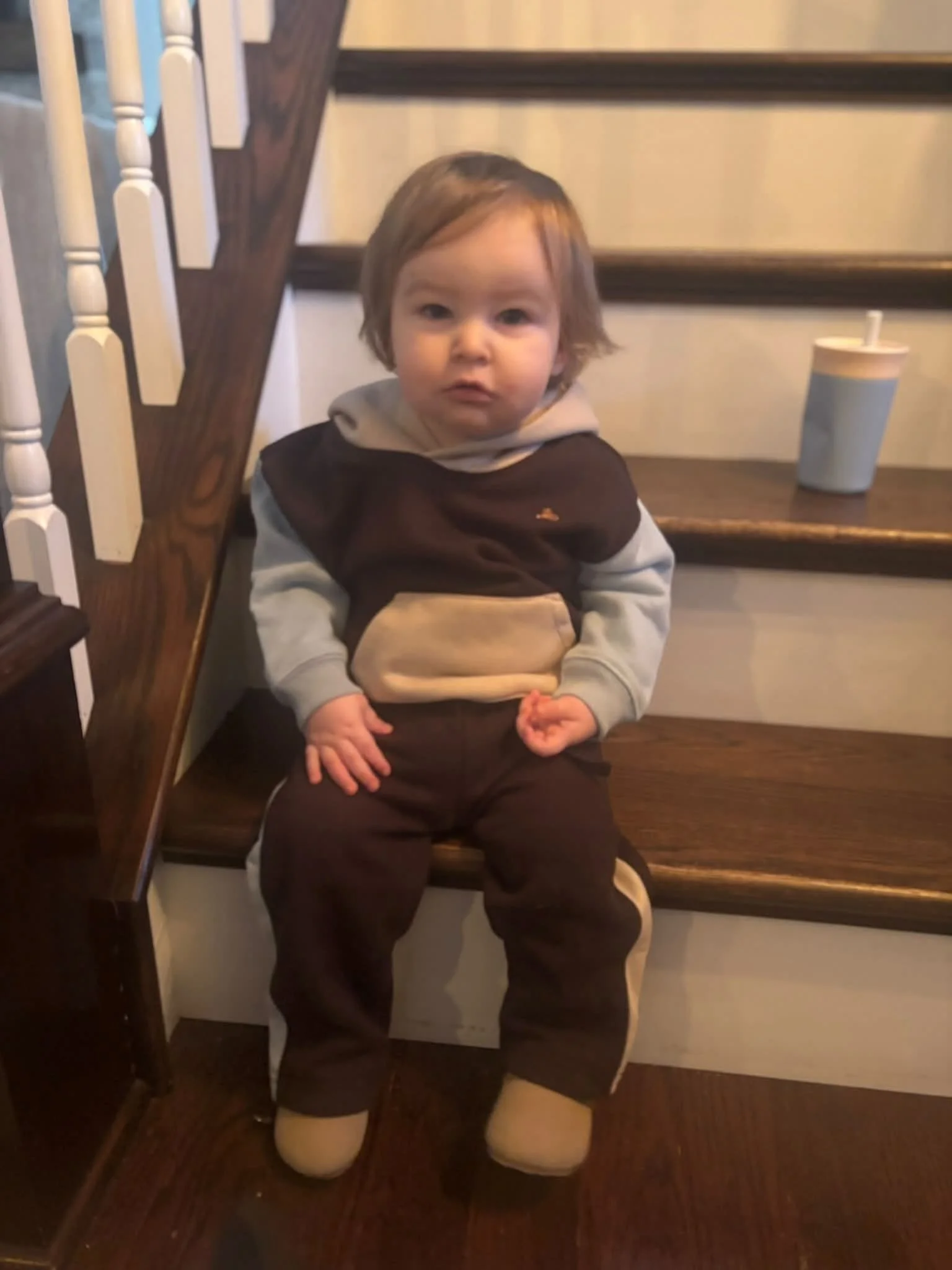 A young child with light brown hair sitting on a wooden staircase, wearing a brown and beige jacket and matching pants, with beige slippers, and a large drink cup on the staircase beside them.