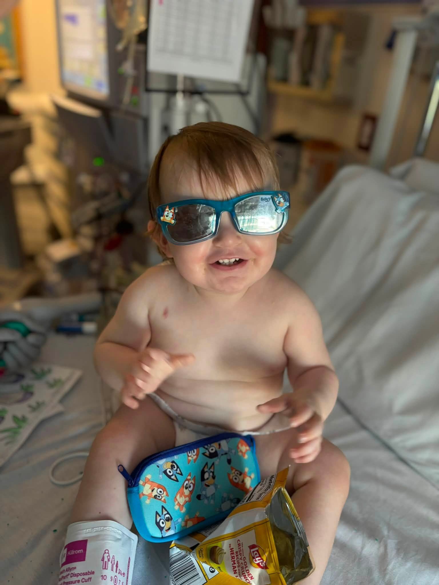 A young child with brown hair wearing funny glasses with cartoon characters is smiling in a hospital or clinic setting. The child is shirtless and sitting on a medical bed. There are medical supplies including a toy and a snack in front of him. The background shows medical monitors, equipment, and books.