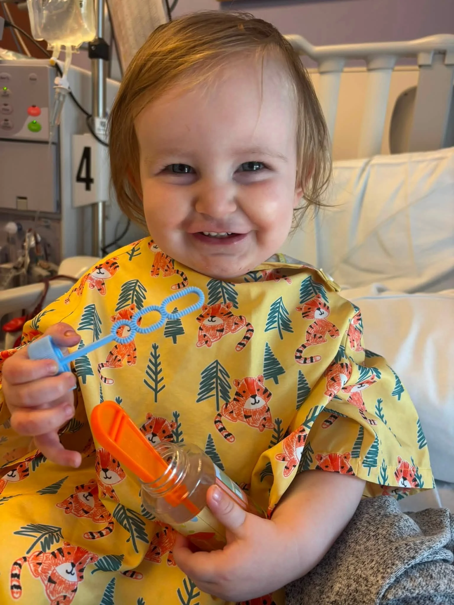 A young child with light brown hair and blue eyes, smiling, seated in a hospital bed wearing a yellow gown with tiger and jungle leaf pattern, holding a plastic bubble wand.