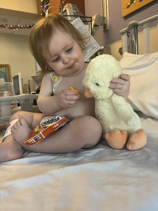 A young girl sitting on a hospital bed, holding a plush duck toy, with a package of Goldfish crackers in her lap.