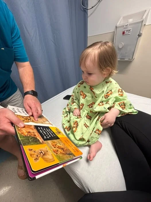 A young child in hospital bed wearing a green animal print gown, sitting upright while looking at a book with pictures of lions and other animals. An adult is holding the book for the child.