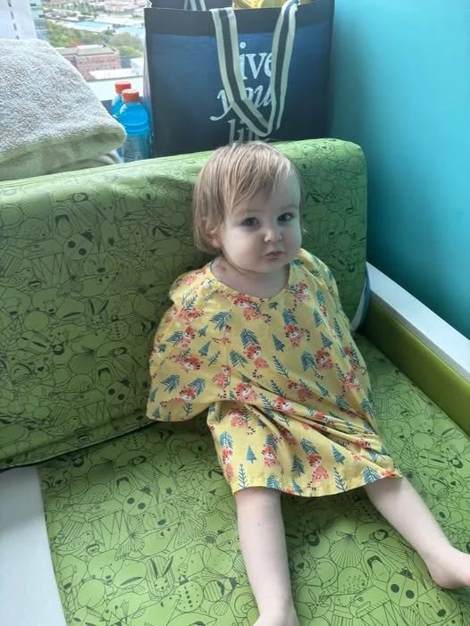 A young boy with short light brown hair and a slight expression of curiosity or confusion sitting on a green patterned seat in a brightly lit indoor space, with a window showing a cityscape in the background in the hospital.