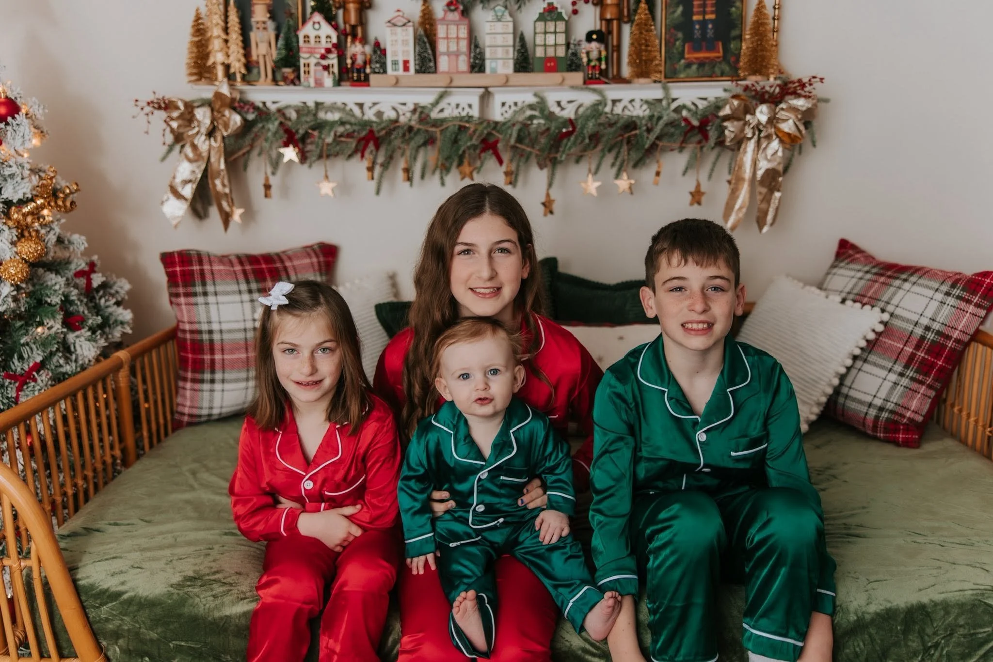 Children dressed in Christmas pajamas sitting on a bed in a festively decorated room with a Christmas tree and holiday decor.