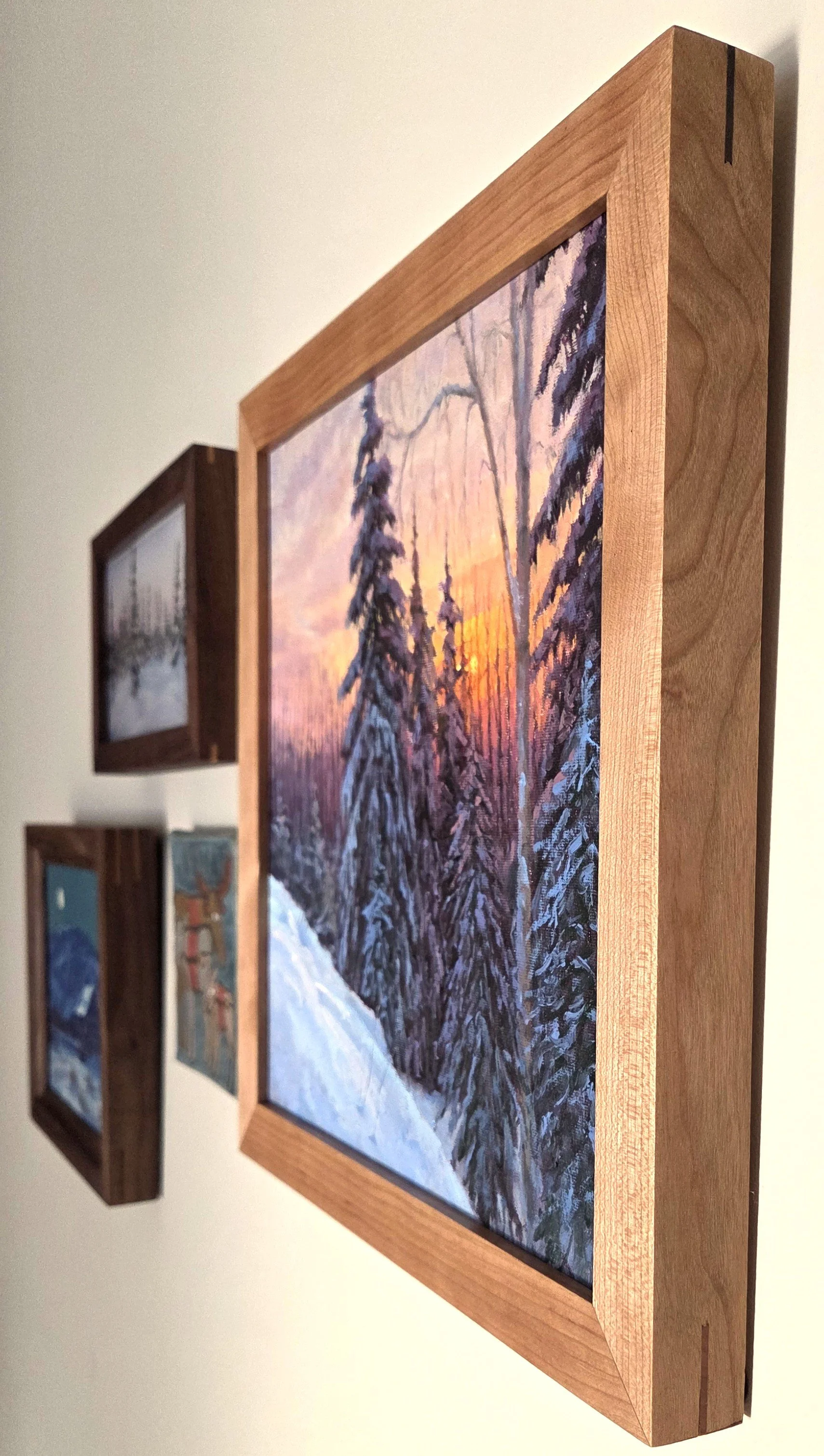 Close-up of framed landscape paintings showing snowy evergreen forests at sunset on a gallery wall