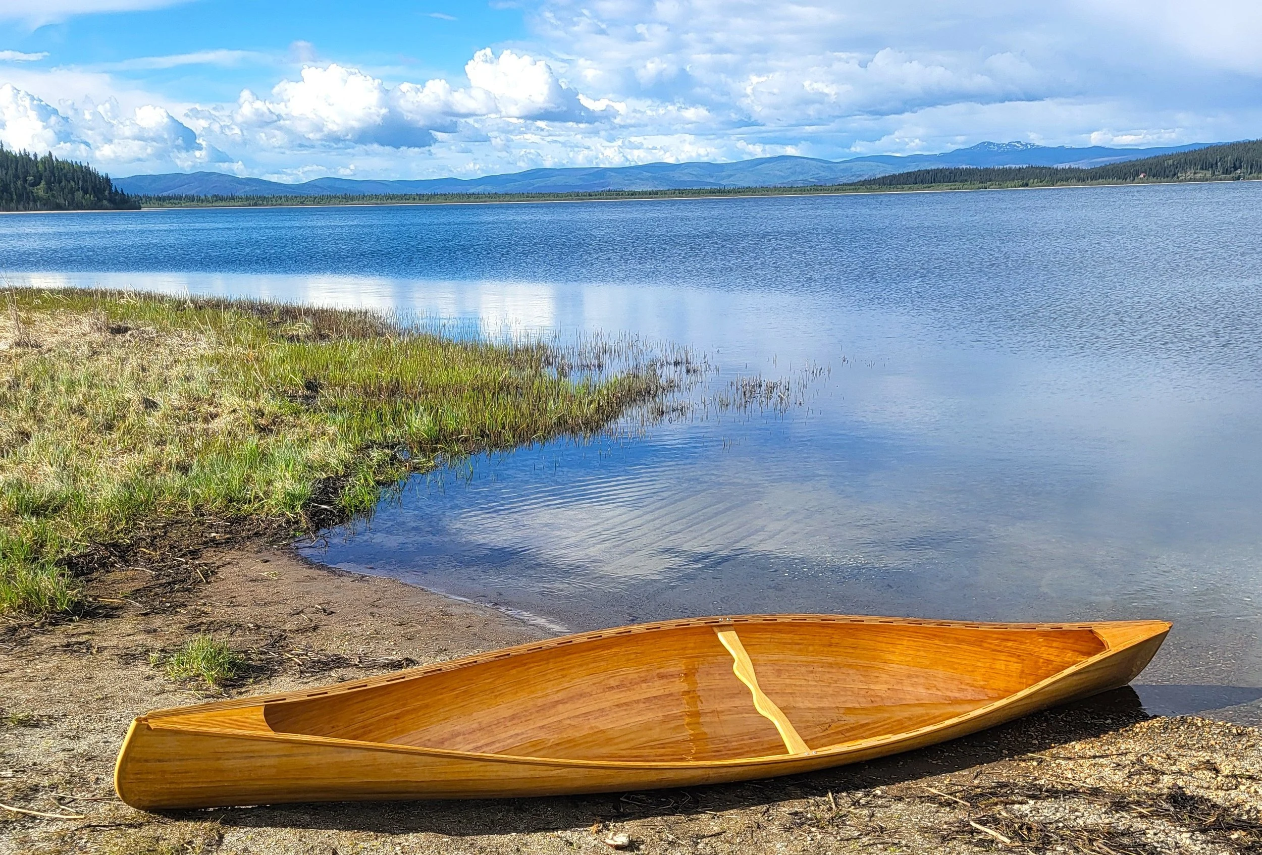 Wooden canoe on a lakeshore with marsh grass, forested mountains, and a cloudy blue sky