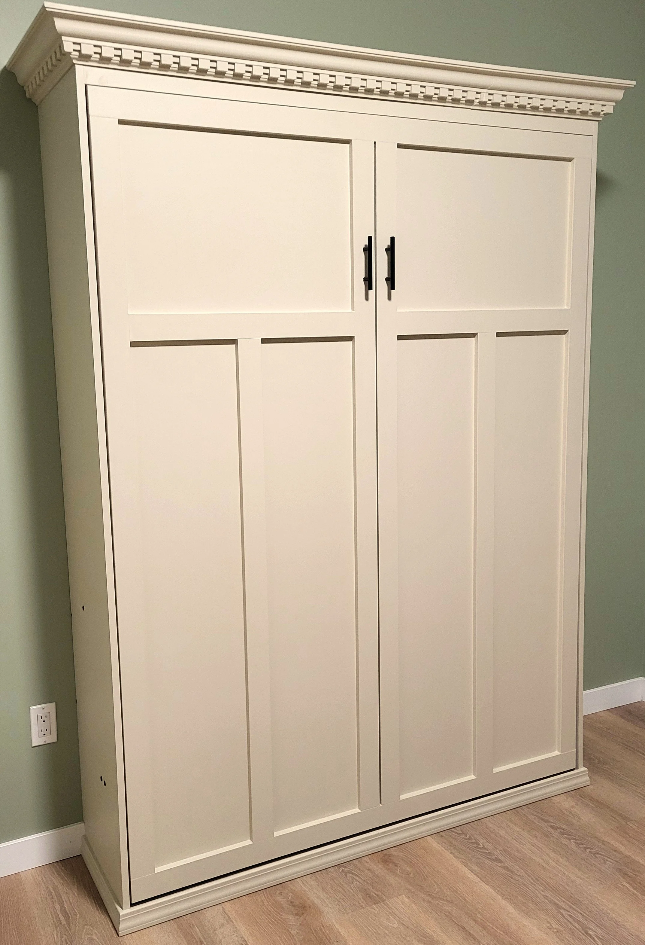 White built-in storage cabinet with paneled double doors, black handles, and decorative crown molding against a green wall.