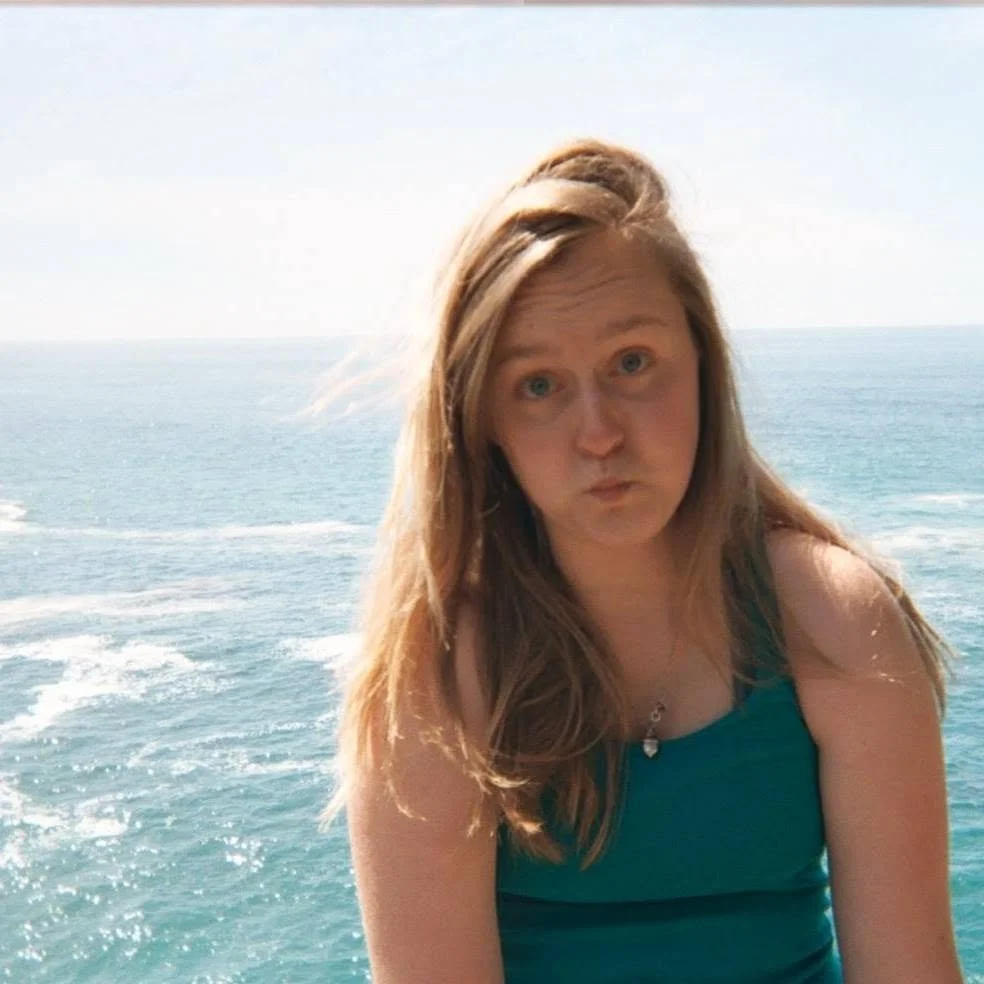 A young woman with long red hair in a teal tank top, sitting outdoors near the ocean on a sunny day.