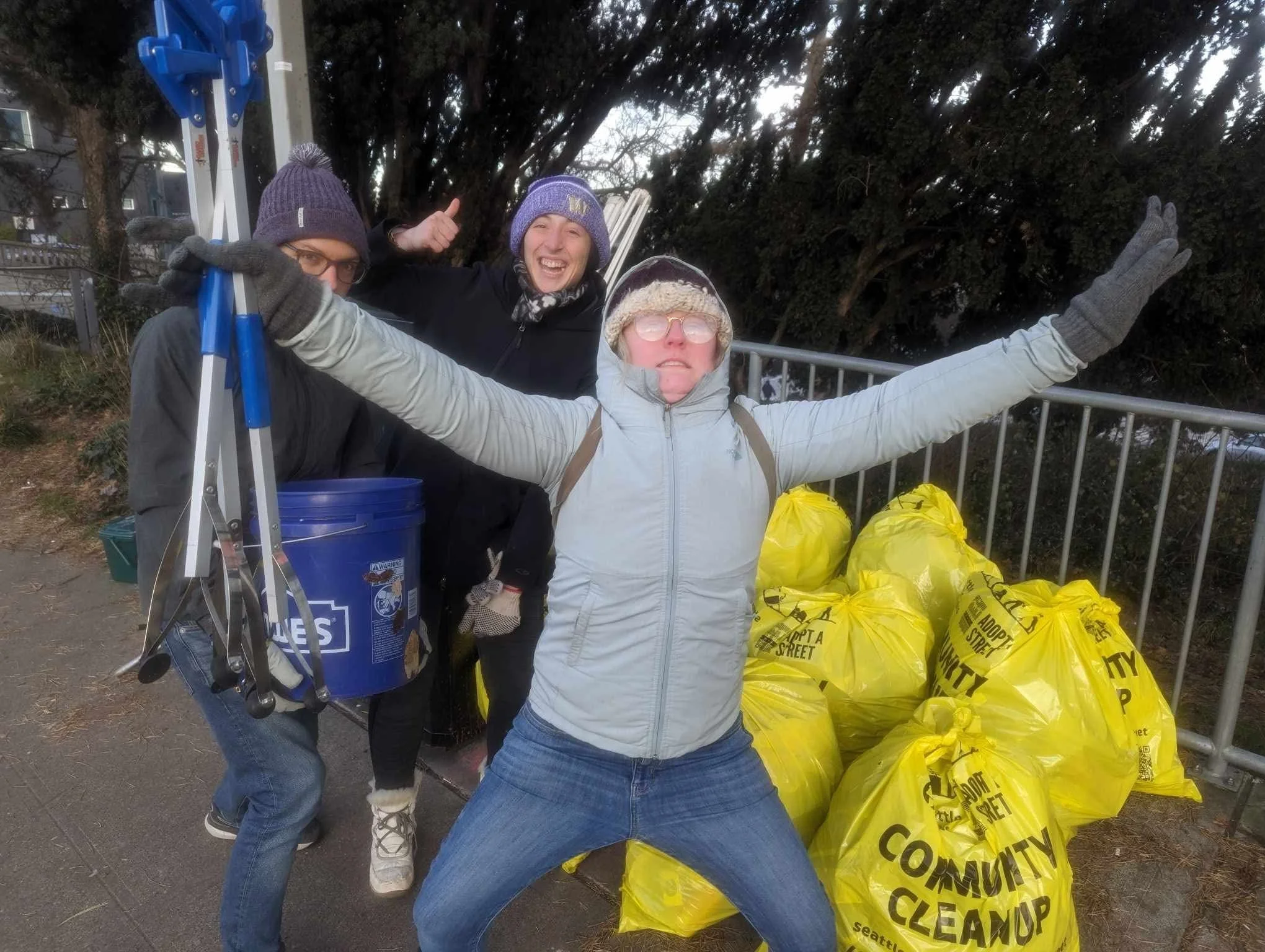 Three people outdoors with large yellow garbage bags labeled 'City Street Clean Up.' One person is holding a blue bucket and ladder, another is giving a thumbs-up, and the third has arms spread wide. All are dressed warmly in hats, gloves, and jackets, and are smiling.