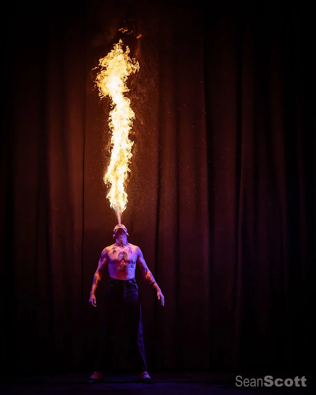 A male fire performer stands on stage with flames coming out of his mouth, directed upward, in front of dark curtains.