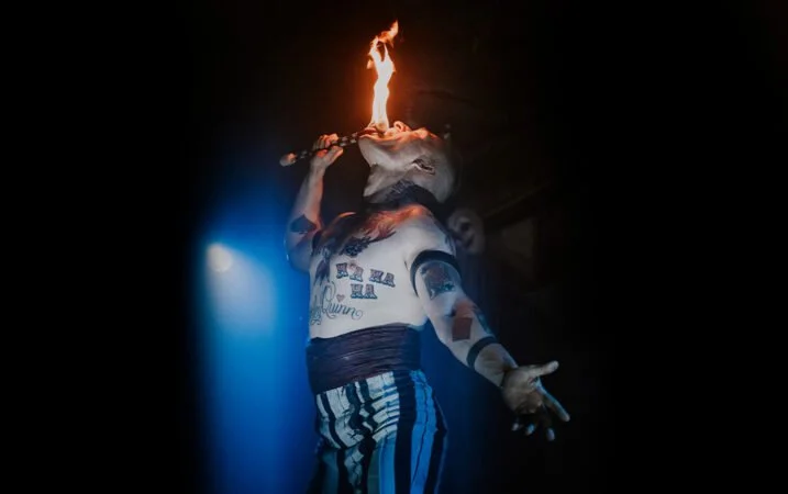 A performer with tattoos and striped pants is holding a flaming object above their mouth during a show.