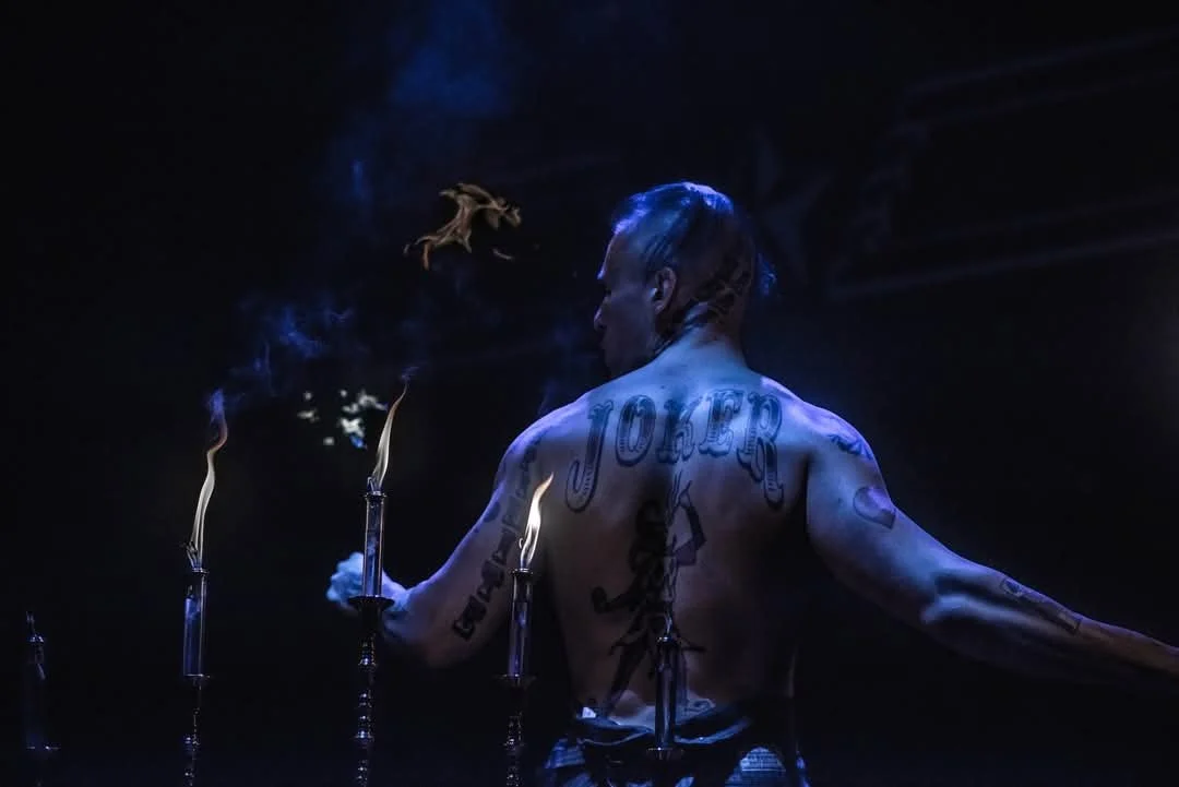 A shirtless person with tattoos, including the words 'JOKER' on their back, standing in a dark environment with candles and a small flame in their hand, with a small fire or creature floating above.