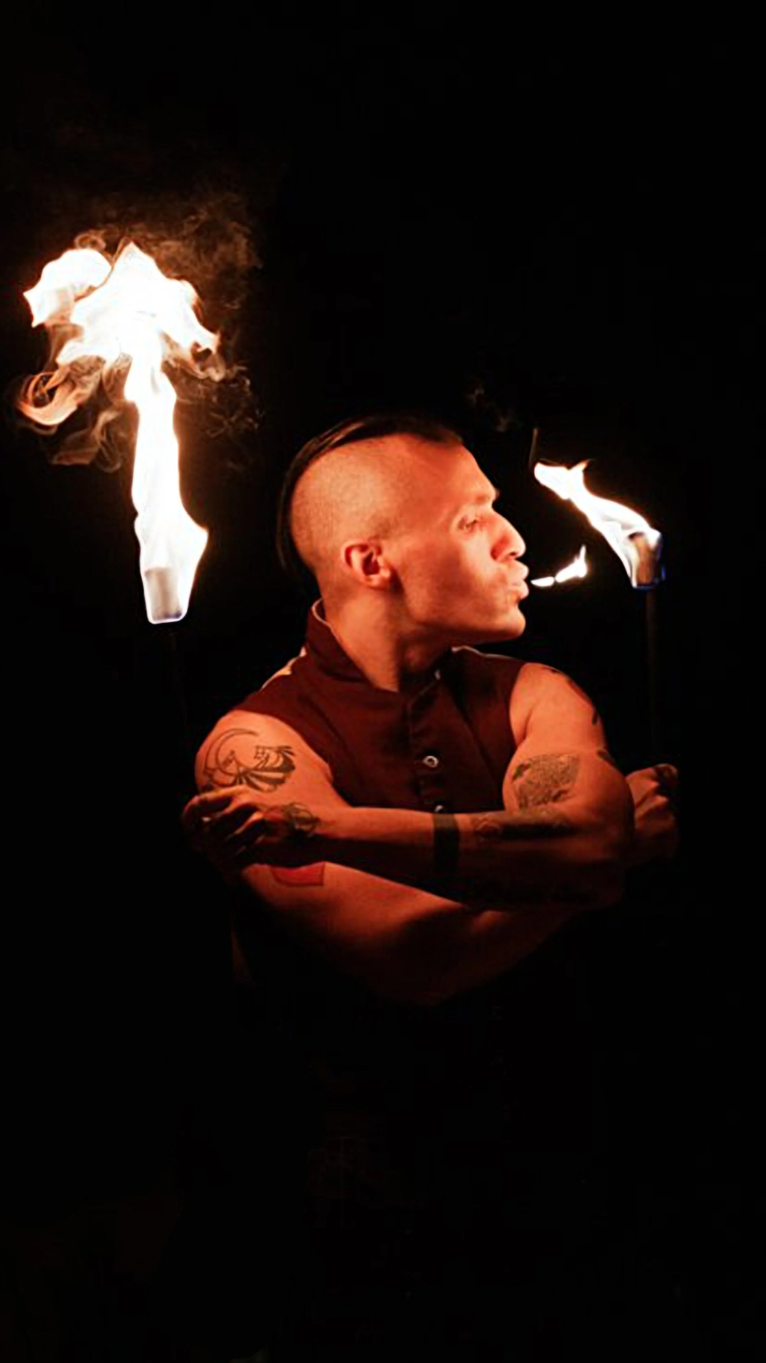 A man with tattoos on his arms and a mohawk hairstyle performing a fire-eating act at night, with flames coming from two torches.