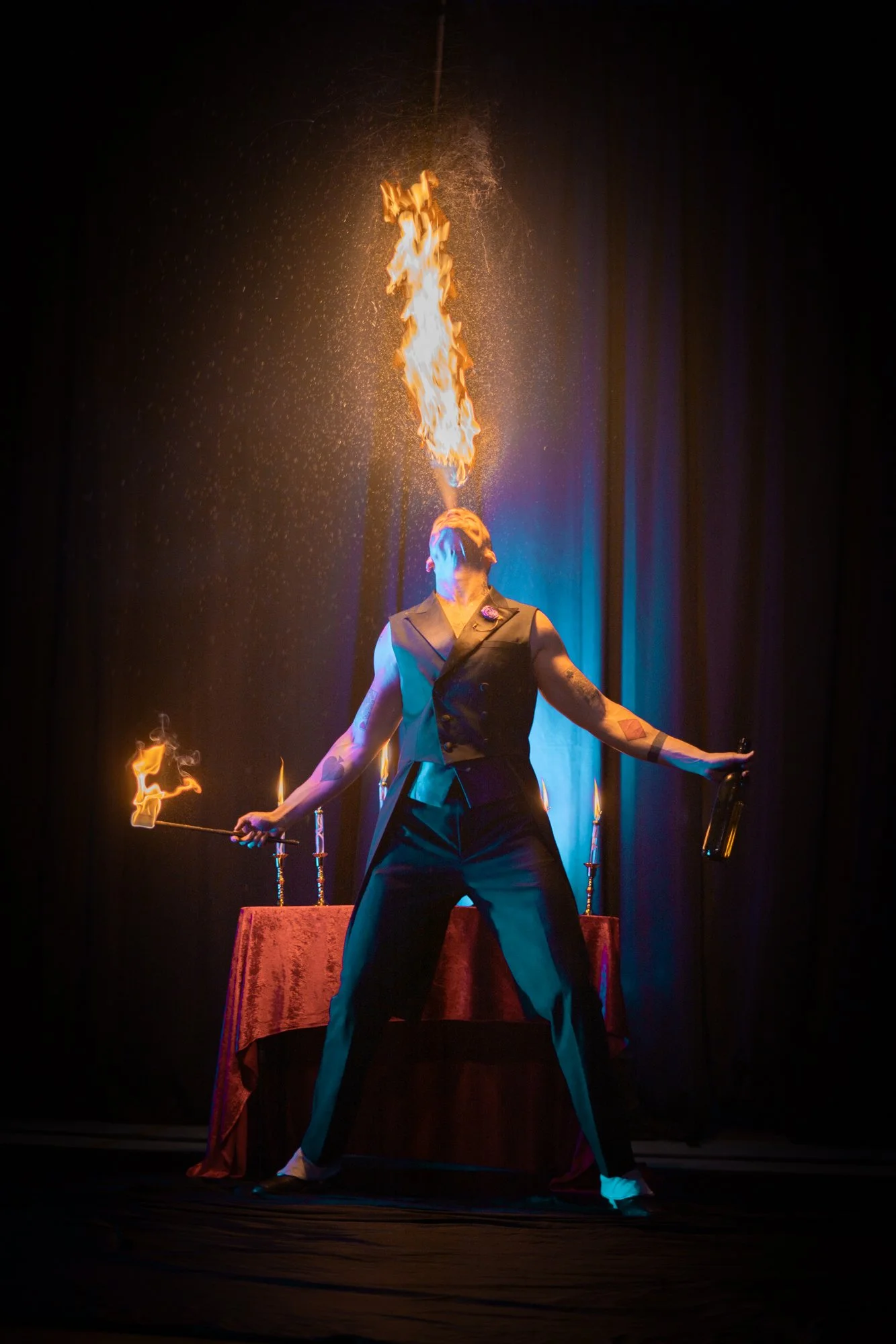 A performer in a black vest and pants stands with arms outstretched on stage, breathing fire, with flames shooting from their mouth and torches in both hands. A red-covered table with candles is behind them, and stage curtains are in the background.