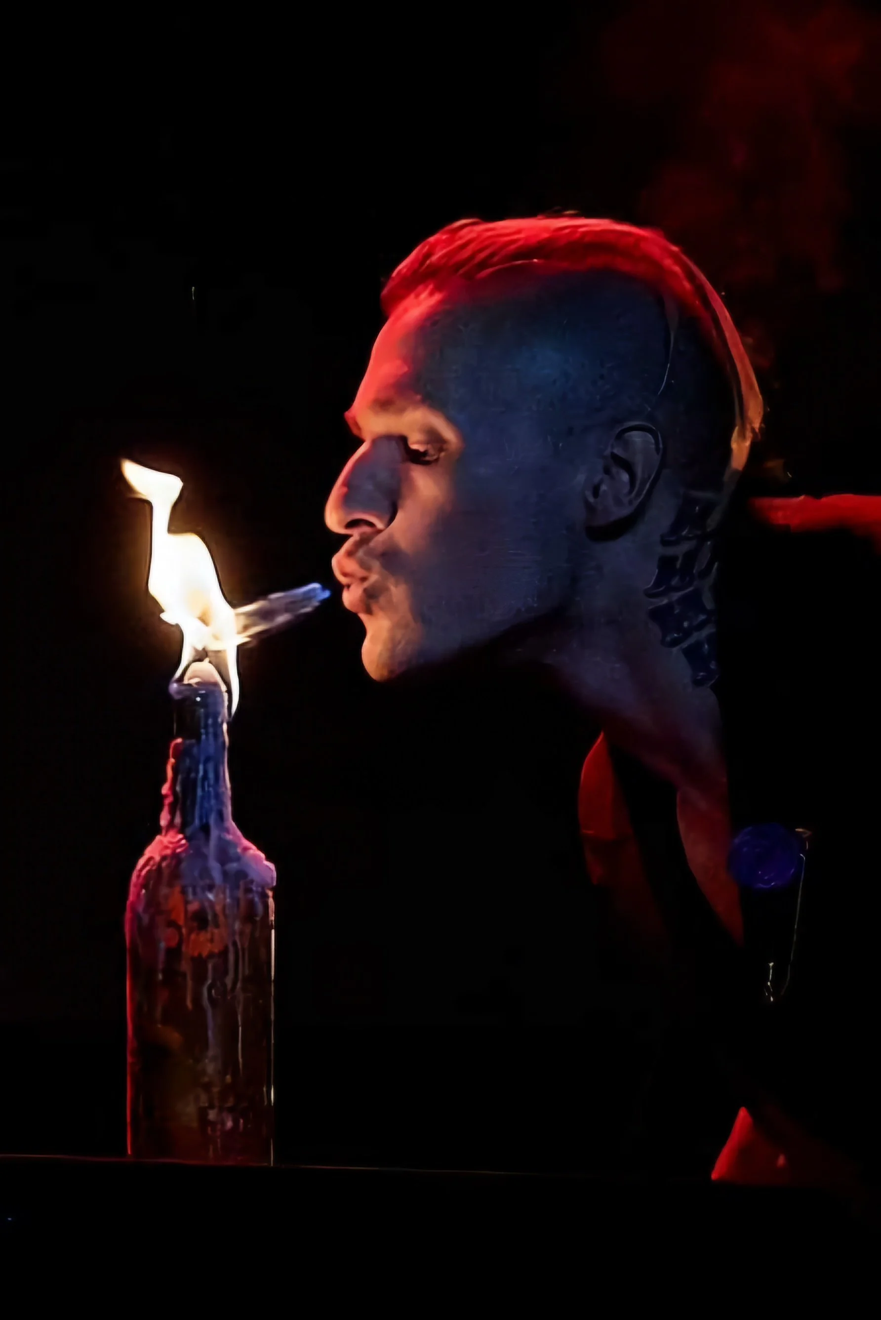 A person with a tattooed head and face is lighting a Molotov cocktail at night. The scene is dark with the flame illuminating the person's face and the bottle.