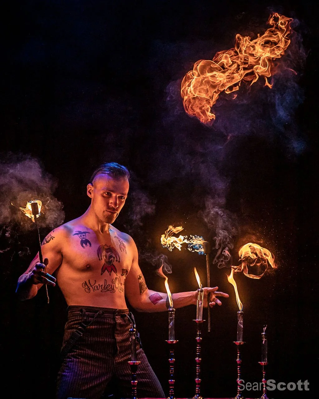 A shirtless male fire performer with tattoos, holding fire torches, manipulates flames and fireballs against a dark background.