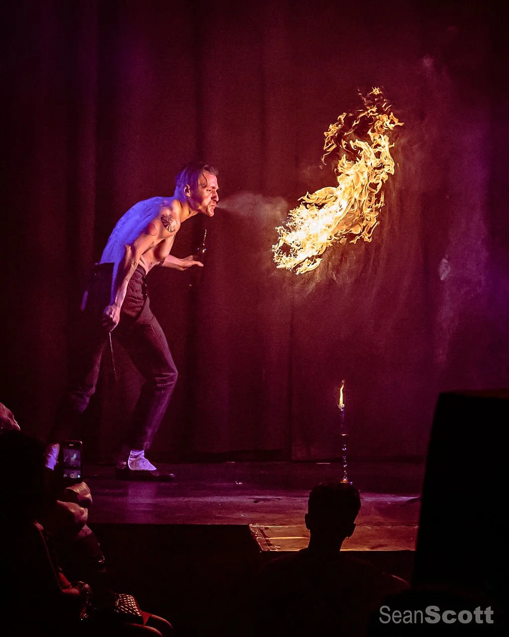 A shirtless male fire performer with tattoos on his arm is standing on stage, blowing a large stream of fire from his mouth. The stage has a dark background.