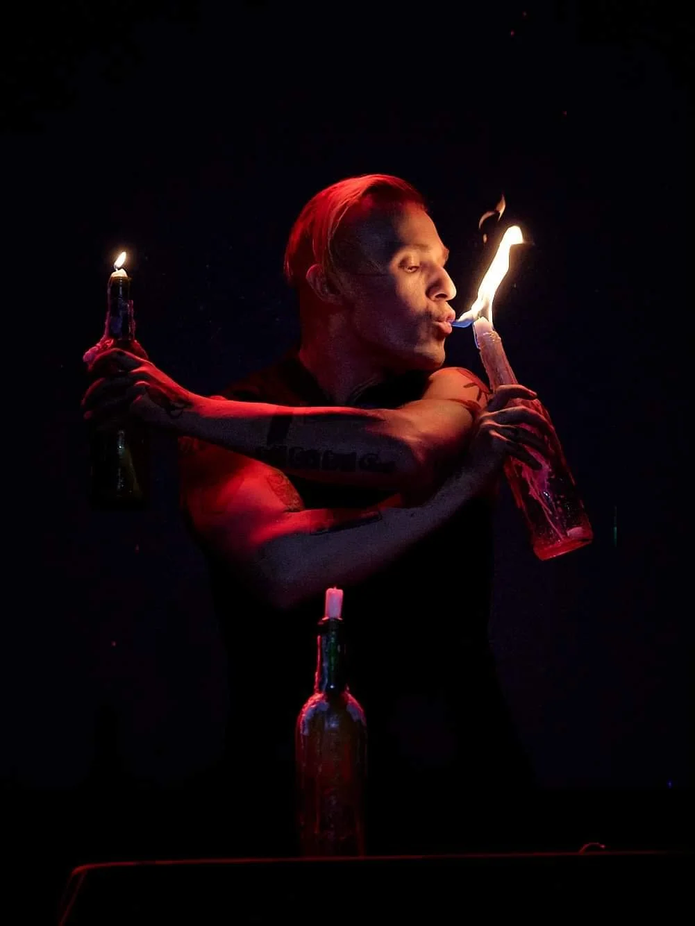 Person with tattooed arms blowing fire from a flame on a bottle, two bottles on either side, dark background, red lighting