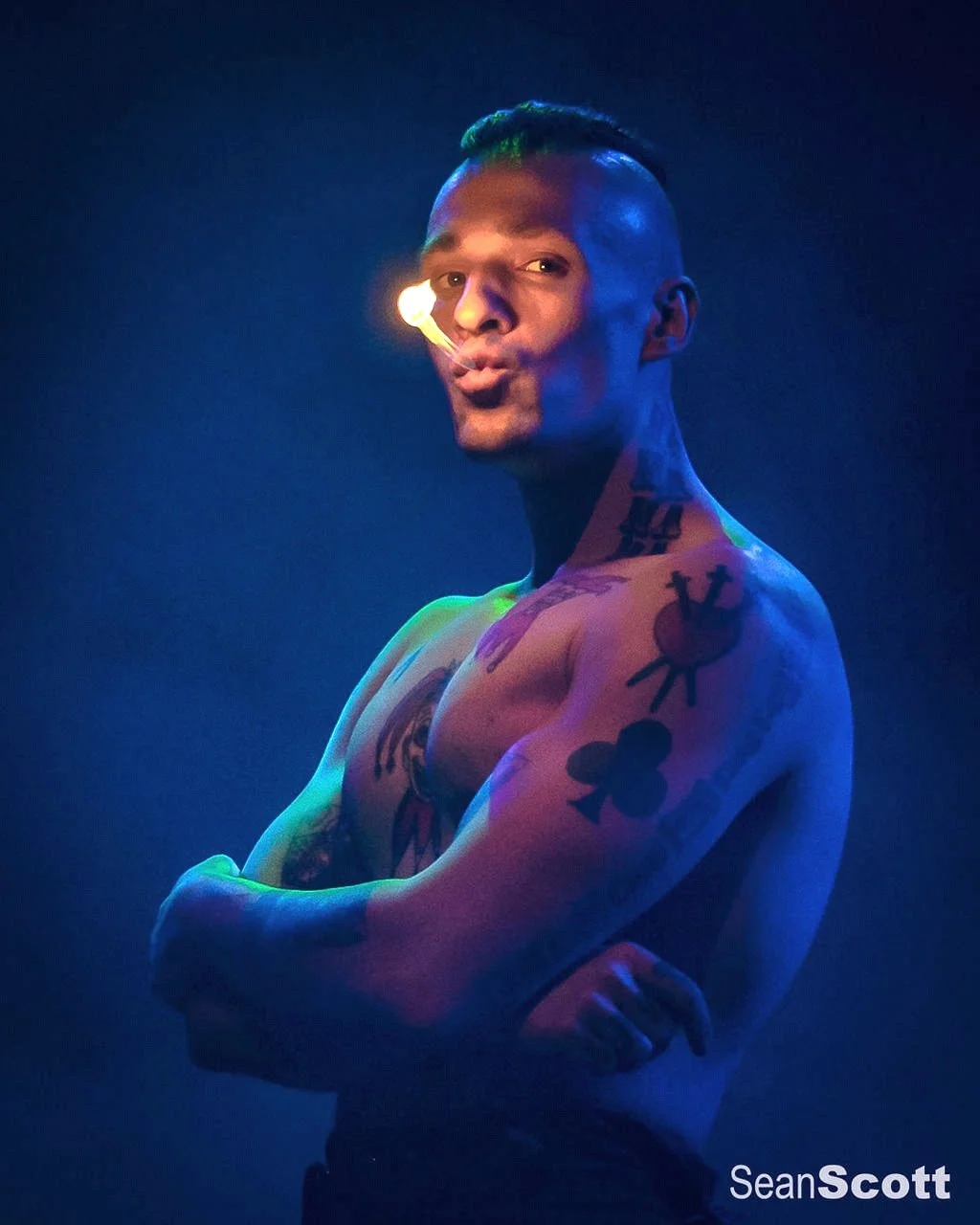 A shirtless man with tattoos on his arm and shoulder, standing with arms crossed, illuminated by dramatic blue and purple lighting, with a casual expression and a light source near his face.