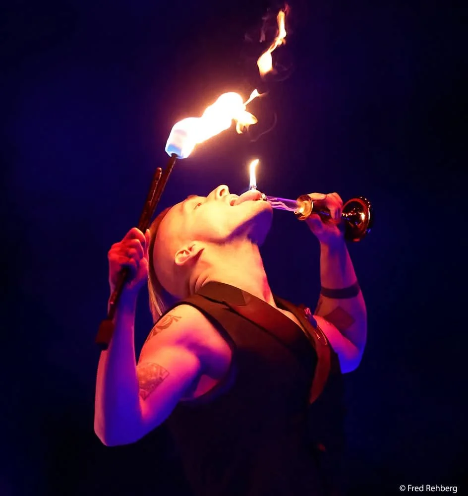 A person performing fire eating, holding a flaming torch in one hand and a drinking vessel in the other, with flames emerging from their mouth against a dark background.