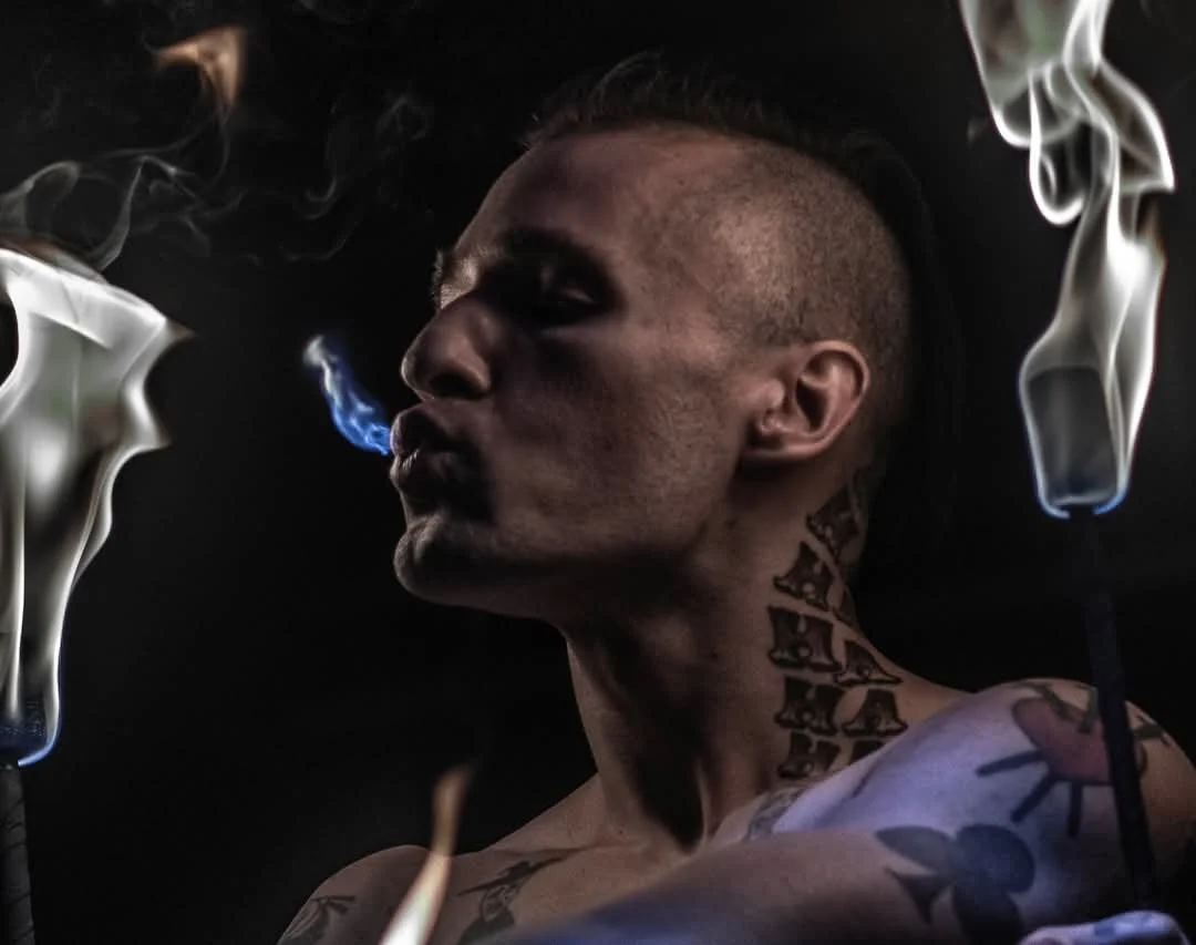 A person with tattoos on their neck and shoulder is smoking, with smoke and flames visible. The background is dark.