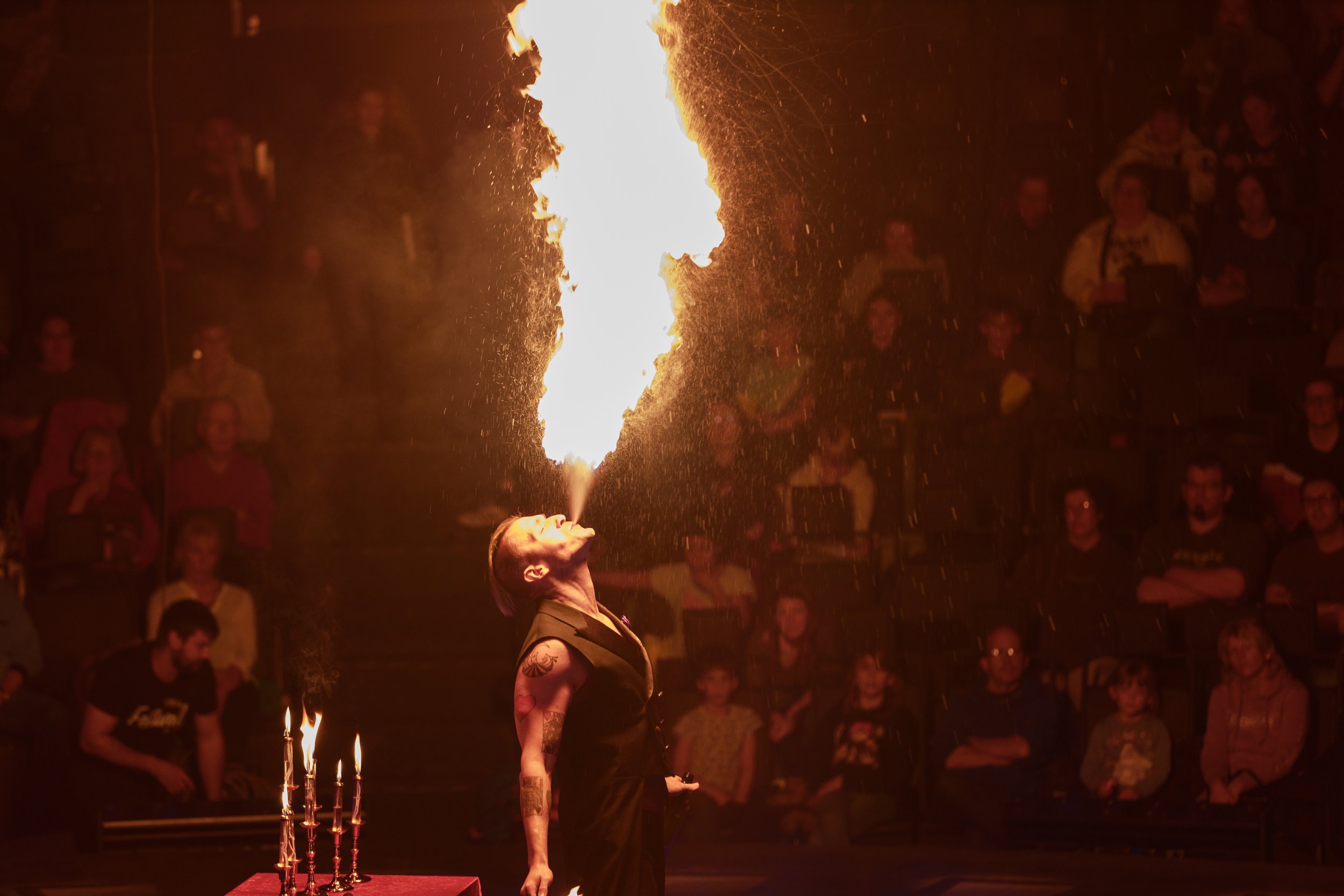 A woman performing a fire-breathing act at a circus or performance event, with a flaming torch in front of her and an audience watching in the background.
