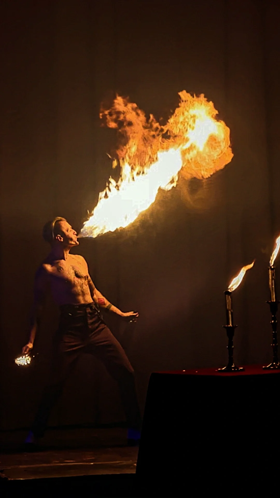 A shirtless male performer with tattoos on his arm, holding a small torch, is breathing fire during a performance. The fire is large and bright, emerging from his mouth toward the ceiling, with two additional sources of fire in front of him. The back
