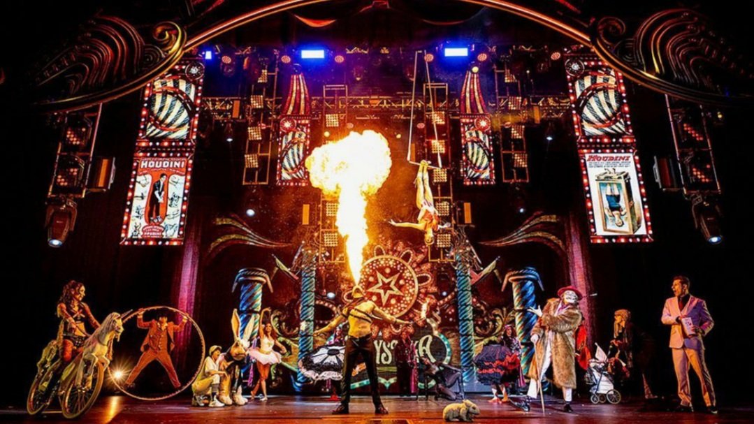 A circus performance featuring acrobats, animals, and fire effects on stage with colorful lights and circus-themed decor.