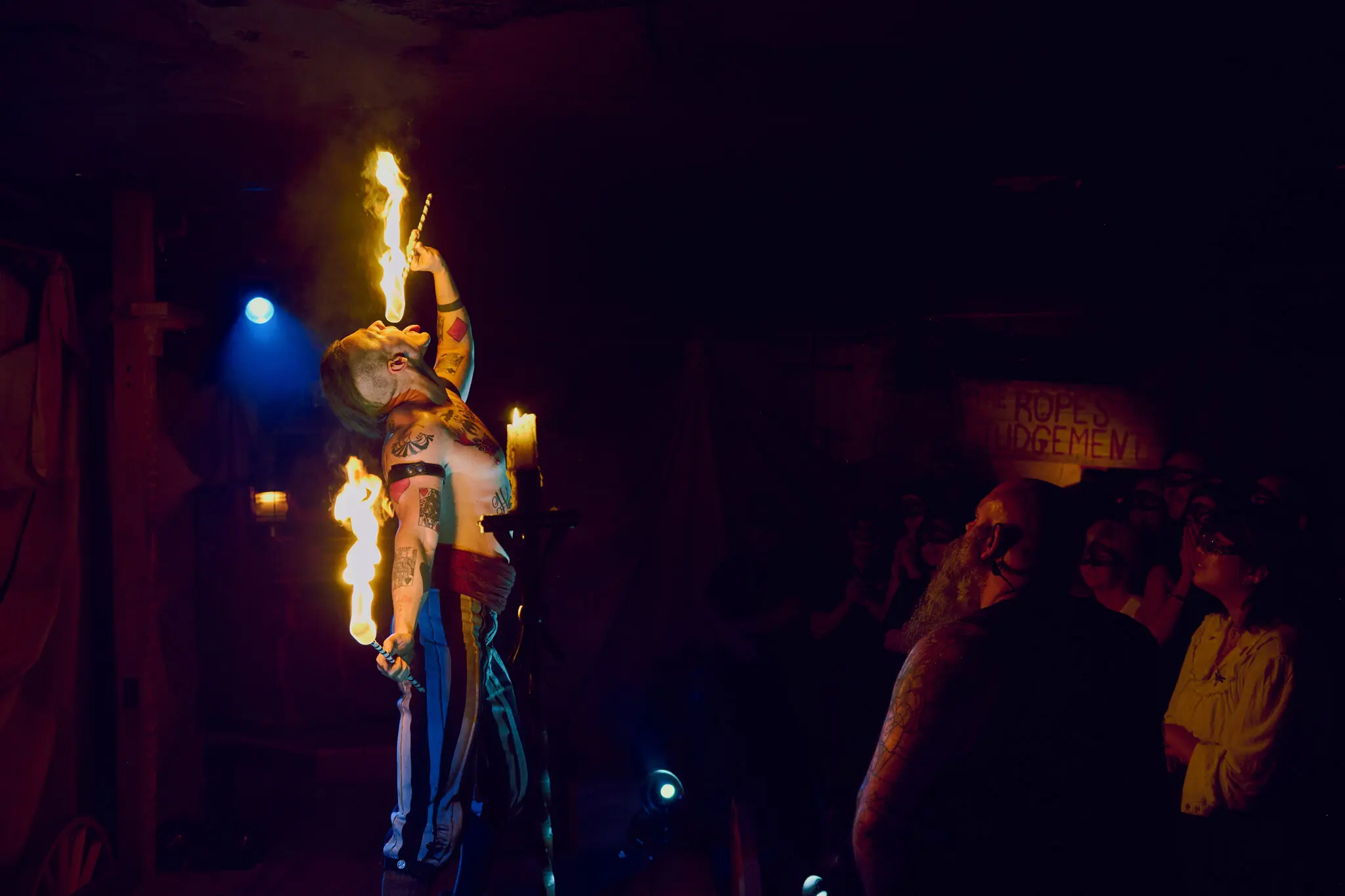 A performer in colorful pants and tattoos breathes fire, holding a torch, during a nighttime show with an audience watching.