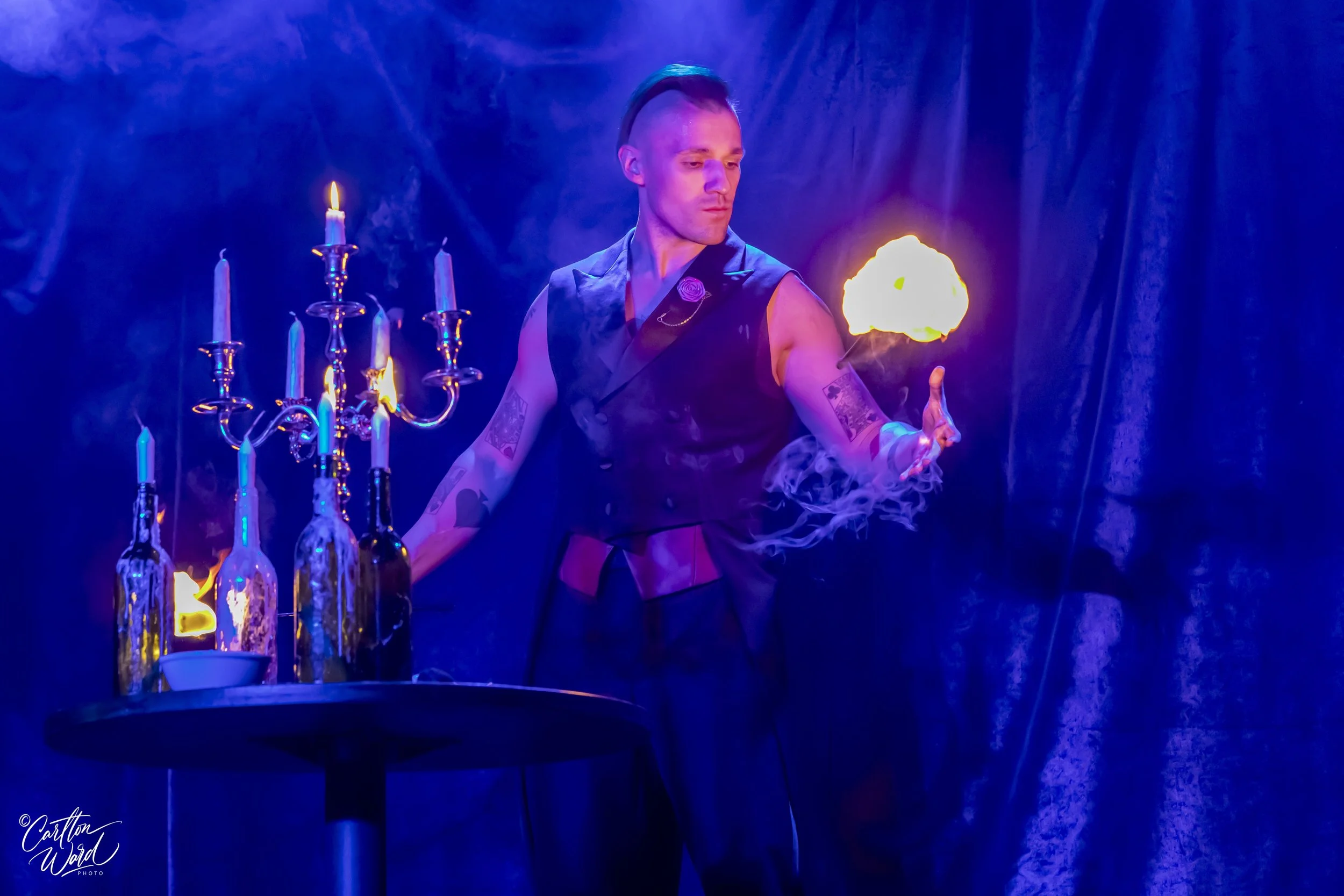 A person with a mohawk hairstyle and tattoos on arms performing a fire act with flames and candle holders, stage or performance setting with dim purple and blue lighting.