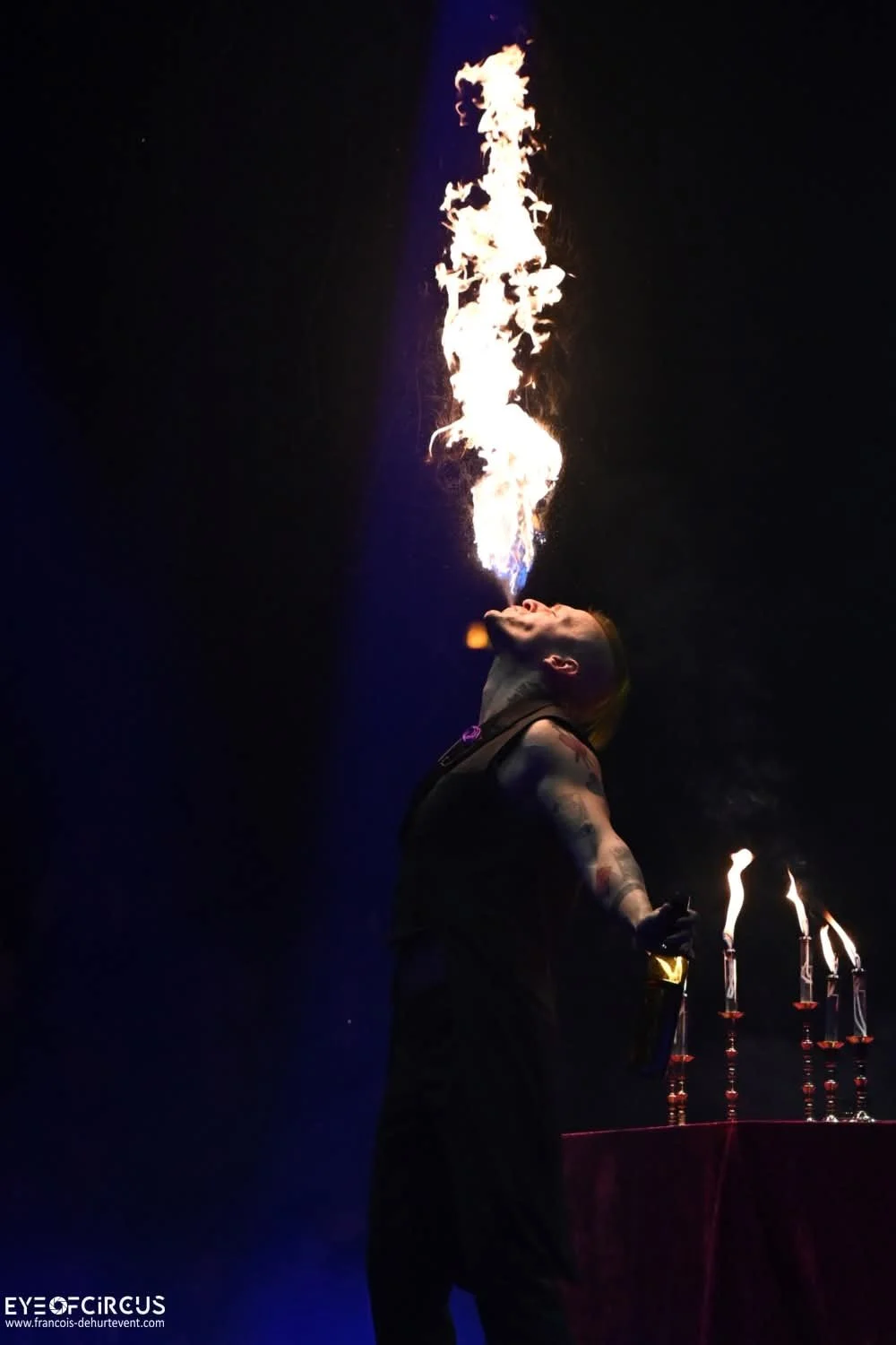 A fire performer is exhaling a large plume of fire into the air, standing in a dark environment with a dark background. The performer has tattoos and is holding a fire torch.