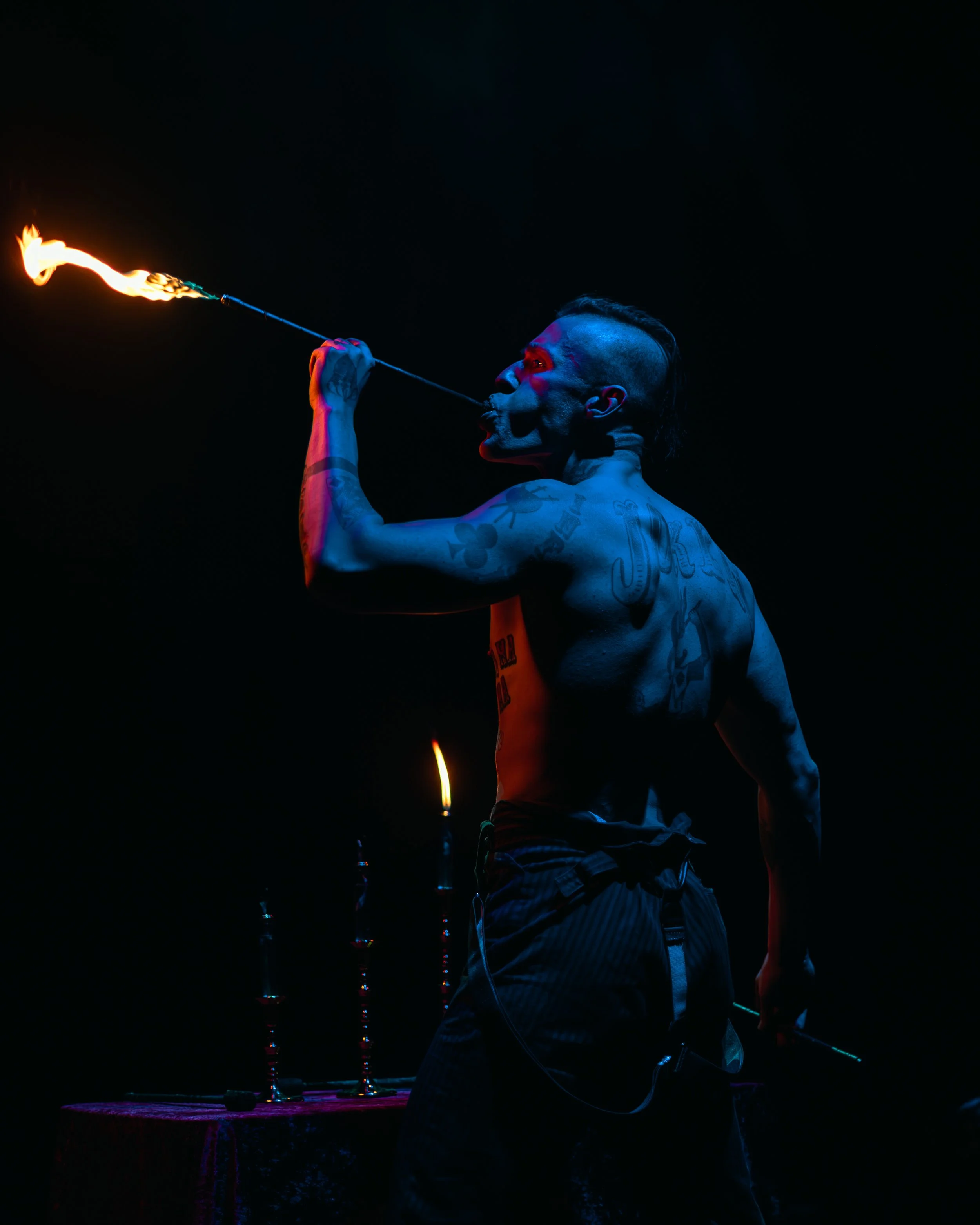 A tattooed man with a mohawk hairstyle is performing a fire-breathing act at night, blowing a flame from a torch he holds, with a dark background and a lit table with multiple fire torches in front of him.