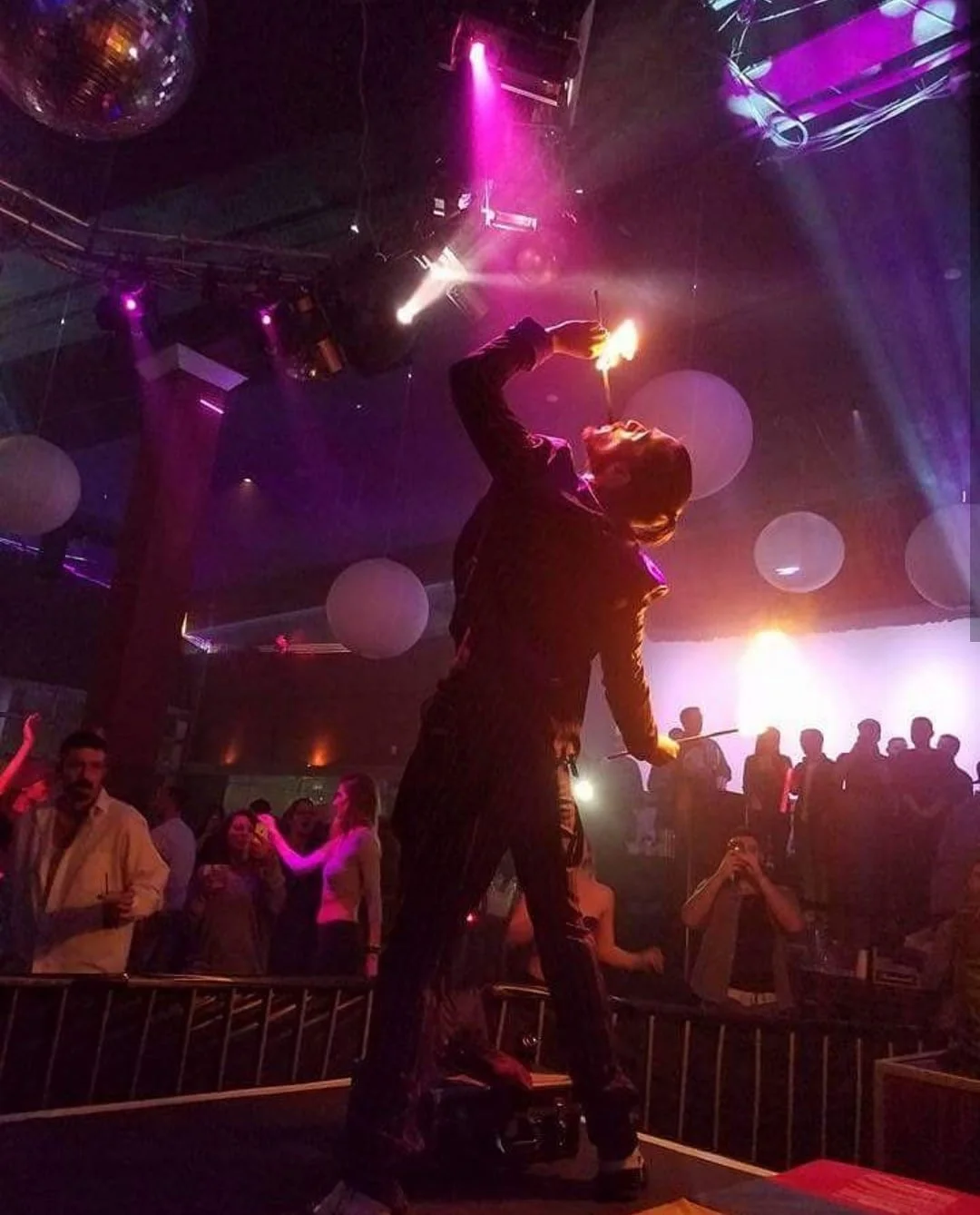 A performer standing on a platform at a nightclub, holding a flame and appearing to be performing a fire act, with colorful lights and dancing crowd in the background.