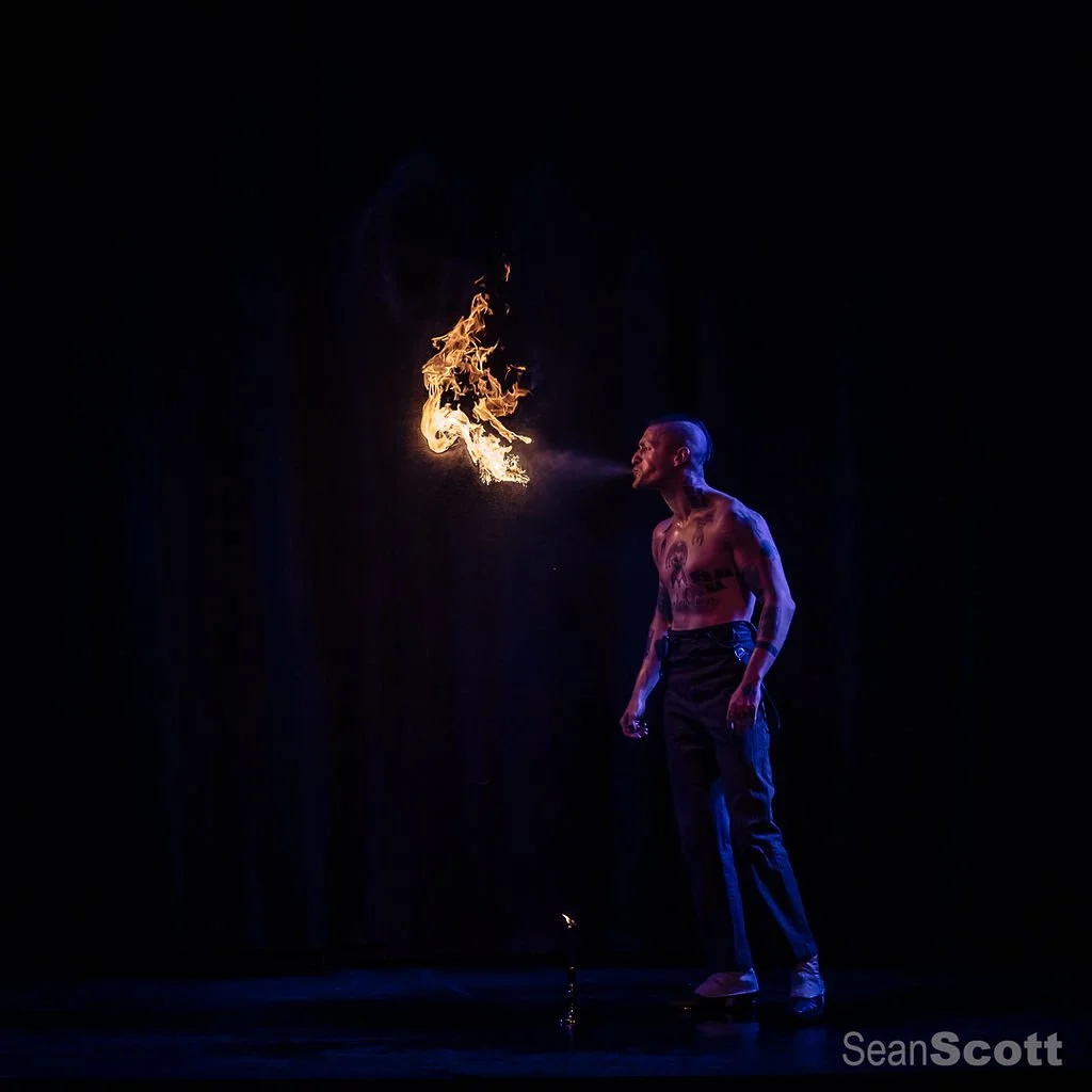 A shirtless male fire performer on stage, blowing fire from his mouth with flames in the air, against a dark background.