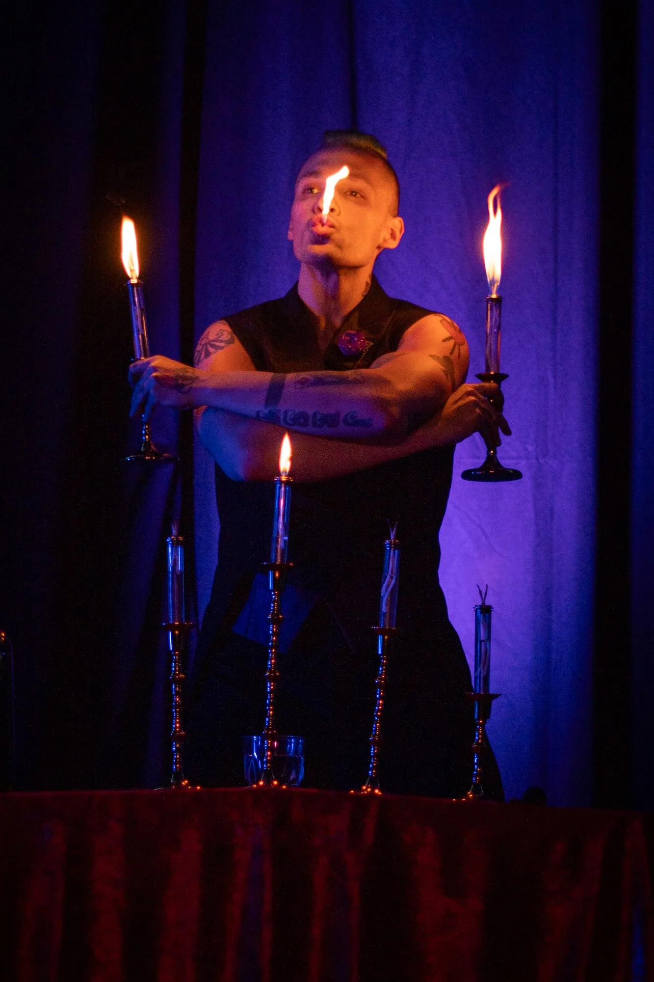 A man with tattoos on his arms, holding three lit candles, performs a fire-eating act on a stage with a dark background.