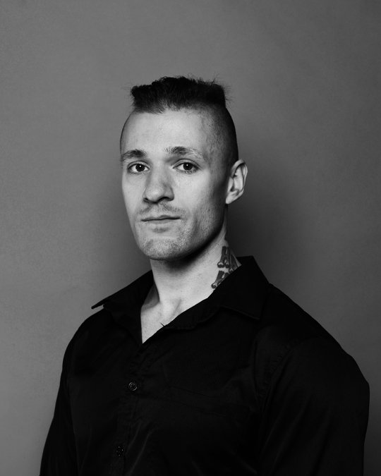 Black and white portrait of a young man with a mohawk hairstyle, wearing a black button-up shirt, standing against a plain background.