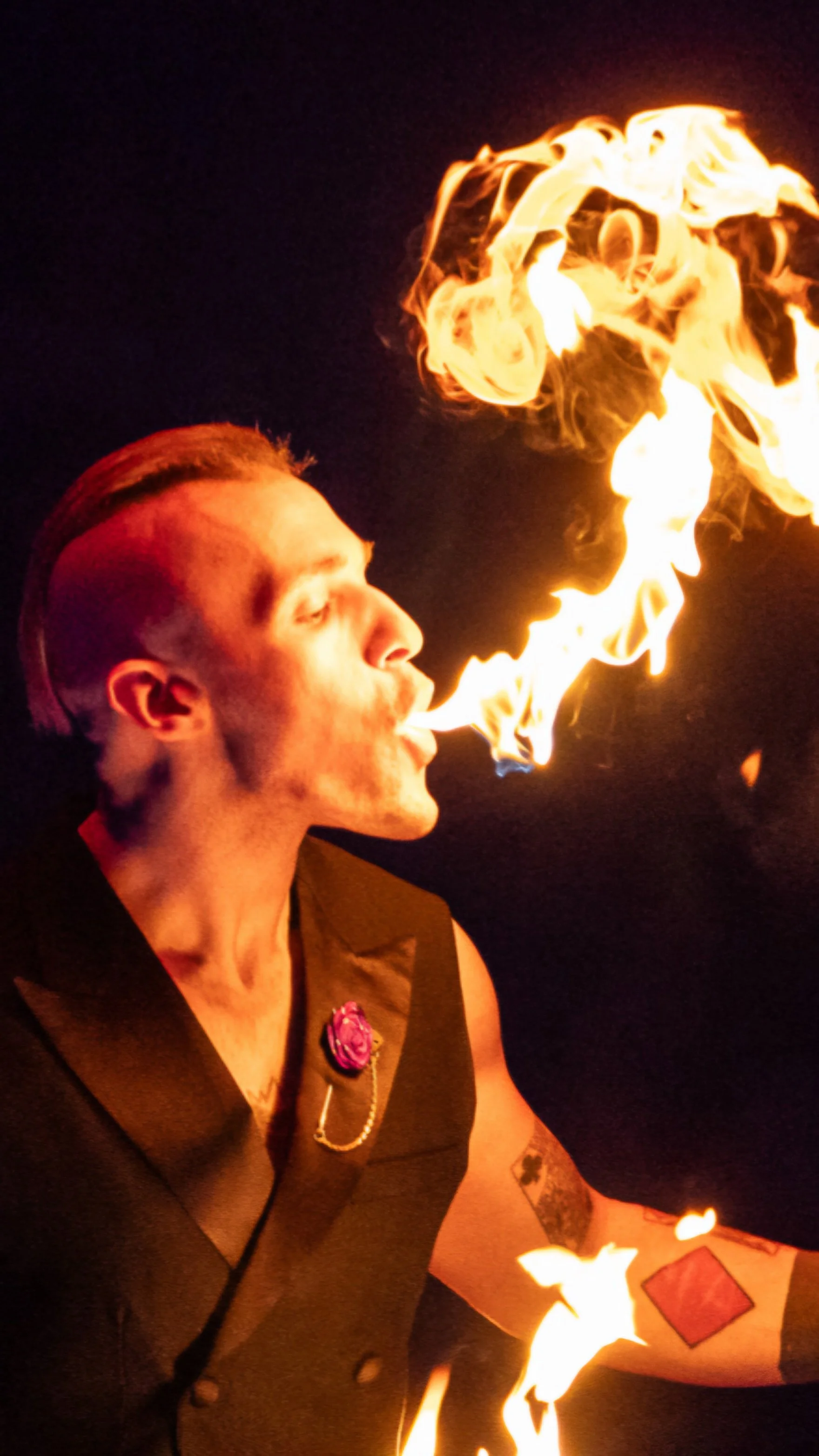 A man with a styled undercut and tattoos on his arm is performing a fire-breathing stunt at night, creating large flames in the air.