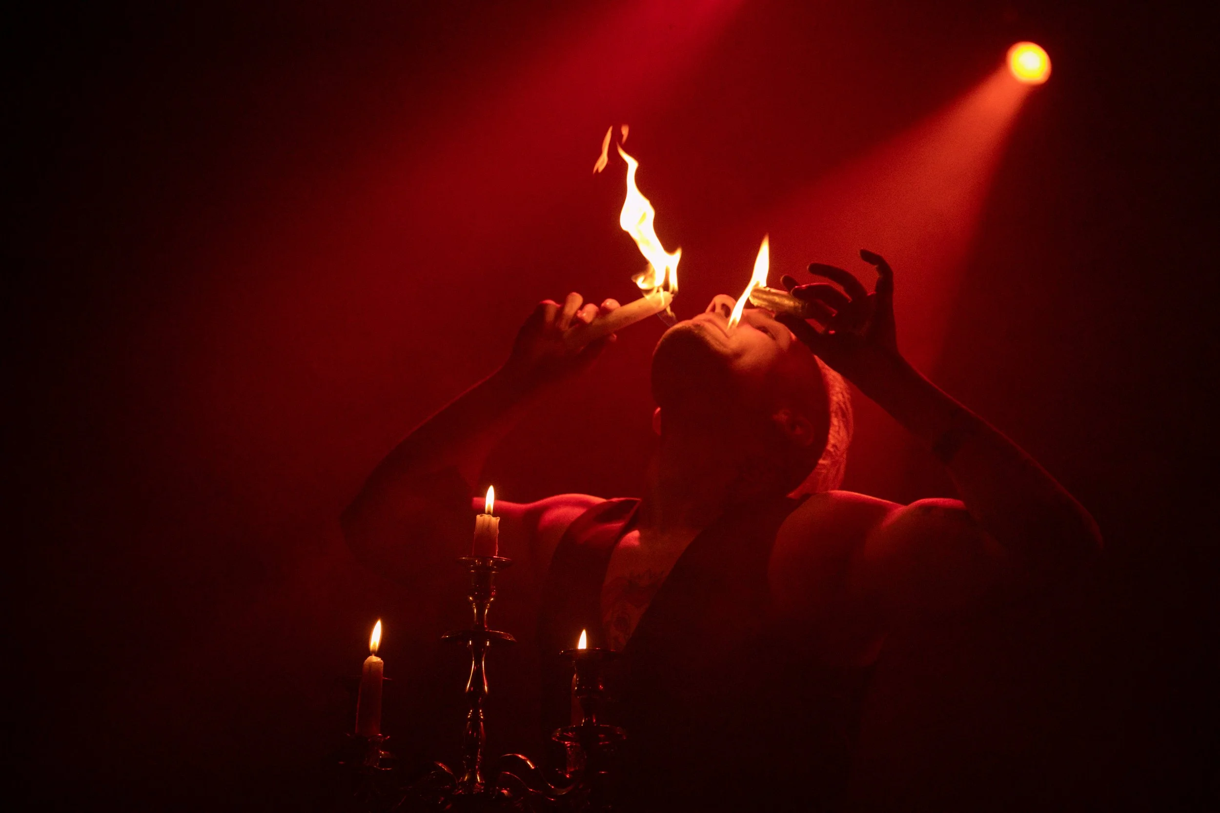 Person performing a fire swallowing act in dim lighting, holding a flaming object to their mouth, with candles and a red spotlight in the background.