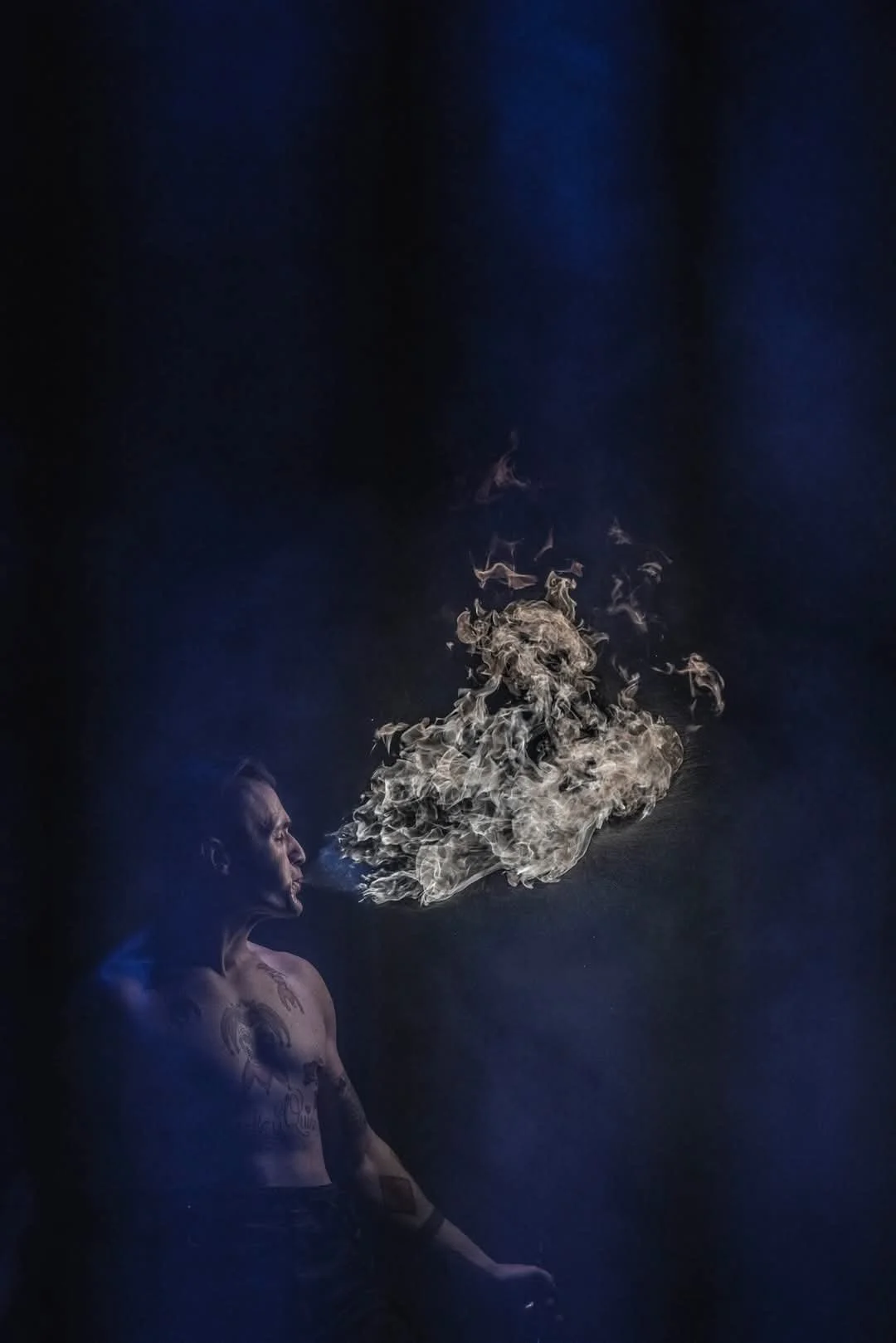 A shirtless man with tattoos on his chest and arm, sitting in a dark space, exhales a large cloud of smoke or vapor, which swirls into the air against a dark background.