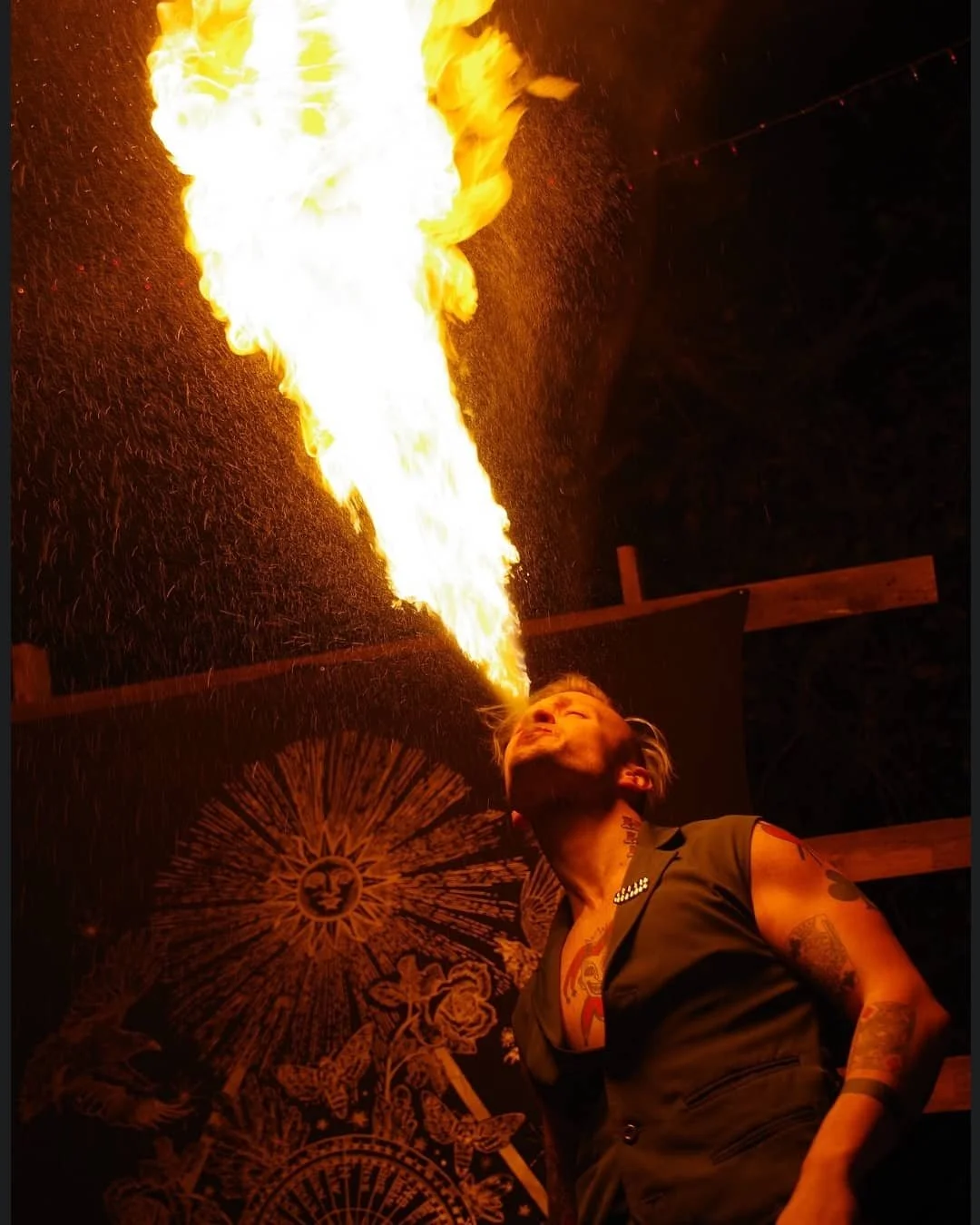 A person with tattoos on their arms and chest performing a fire breathing stunt at night. They are standing under a painted ceiling or wall, and a large burst of fire is erupting from their mouth into the air.