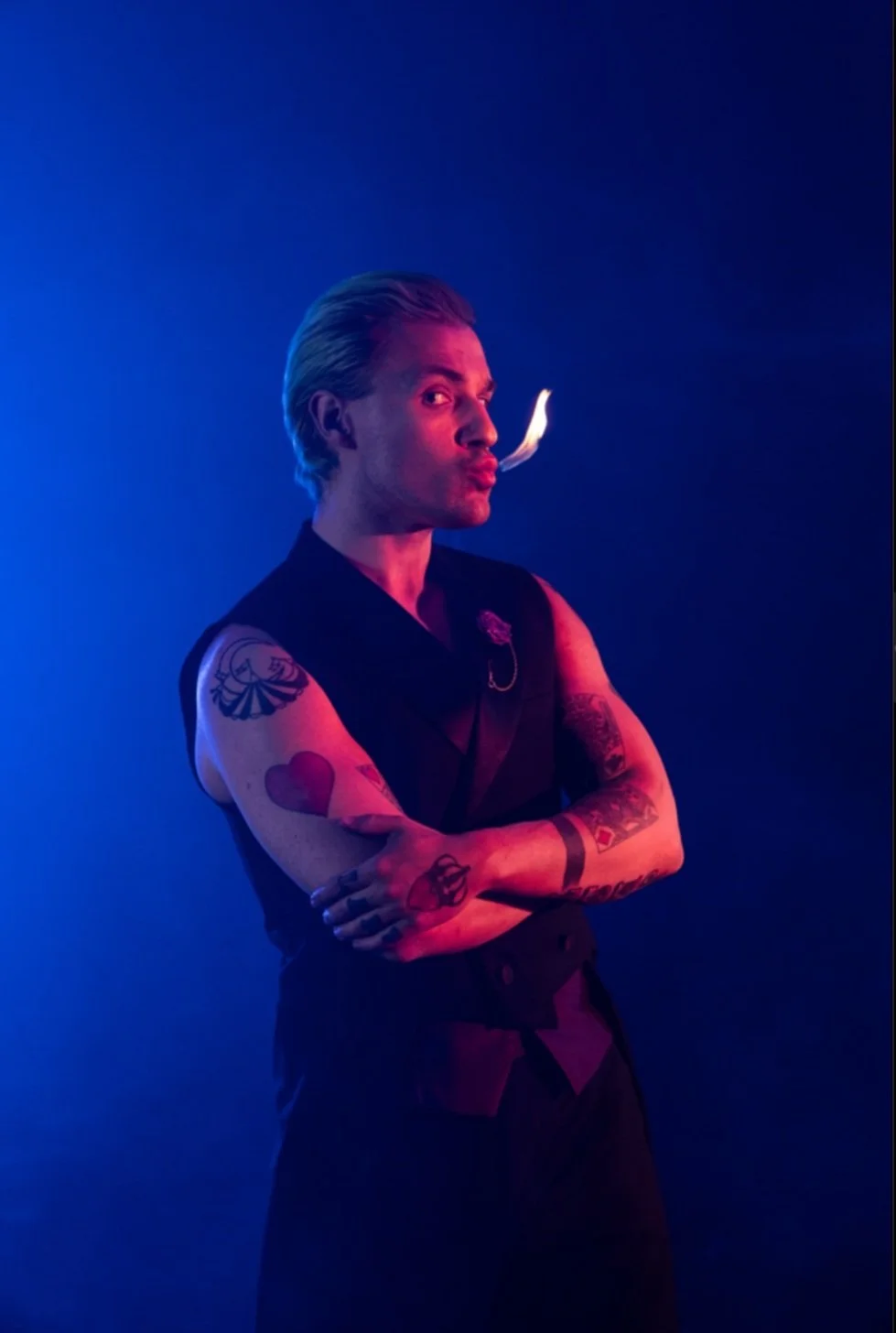 A man with platinum blonde hair, arms crossed, breathing out smoke, in front of a dark blue background with dramatic lighting.
