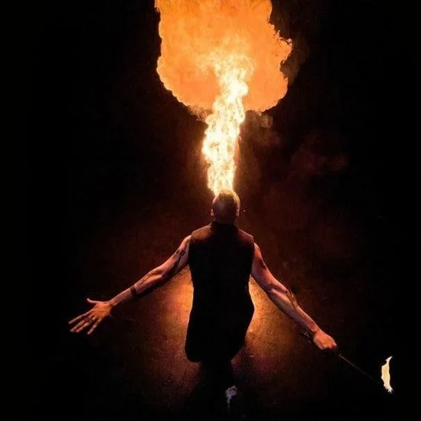 Performer in a dark setting holding a flaming torch in their mouth, with fire coming out, and arms extended outward.