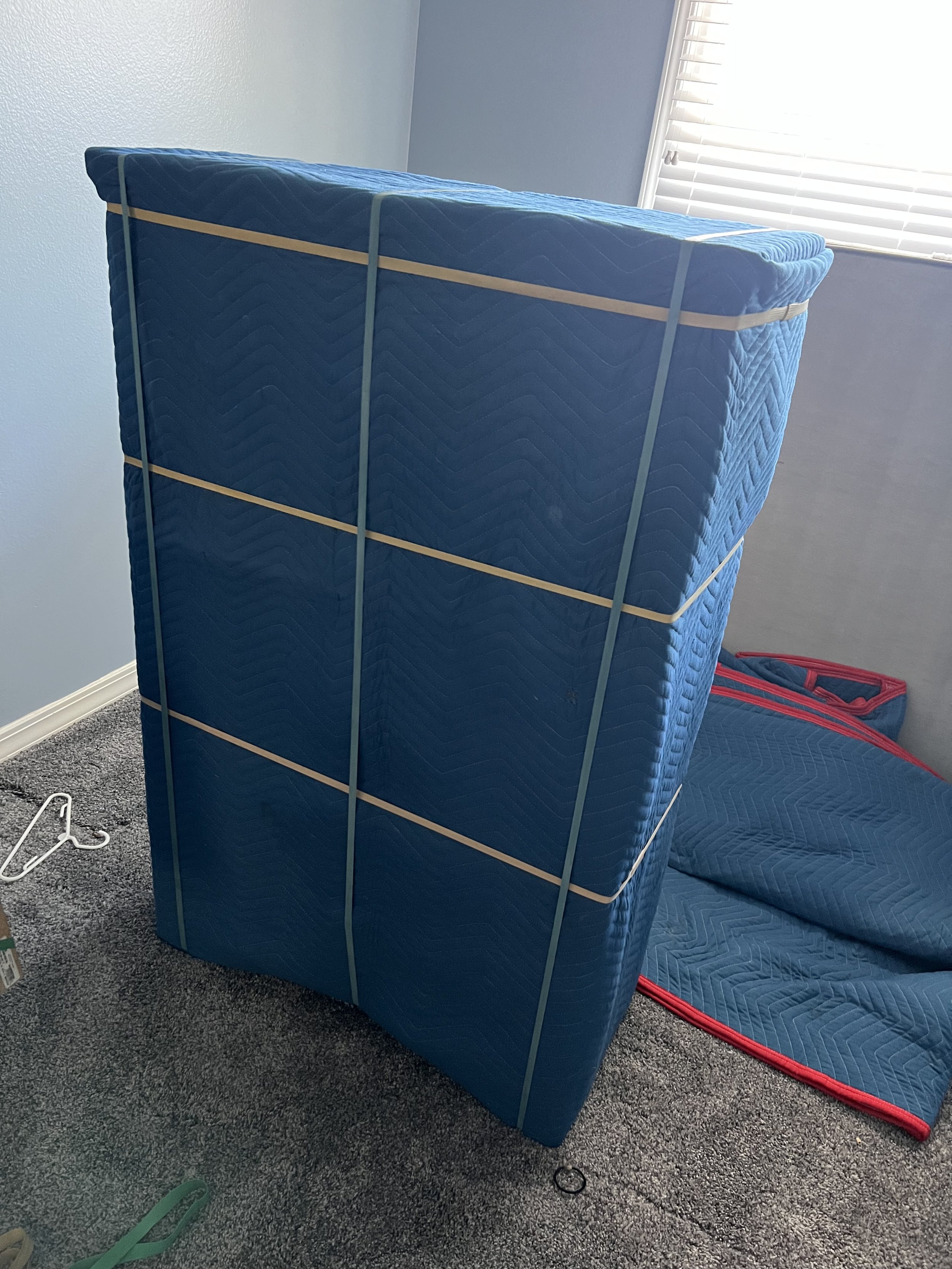 A blue mattress with a quilted pattern standing upright against a wall, secured with green straps, with a blue padded moving blanket on the floor nearby, in a room with gray carpet and light blue walls, and a window with white blinds in the background.