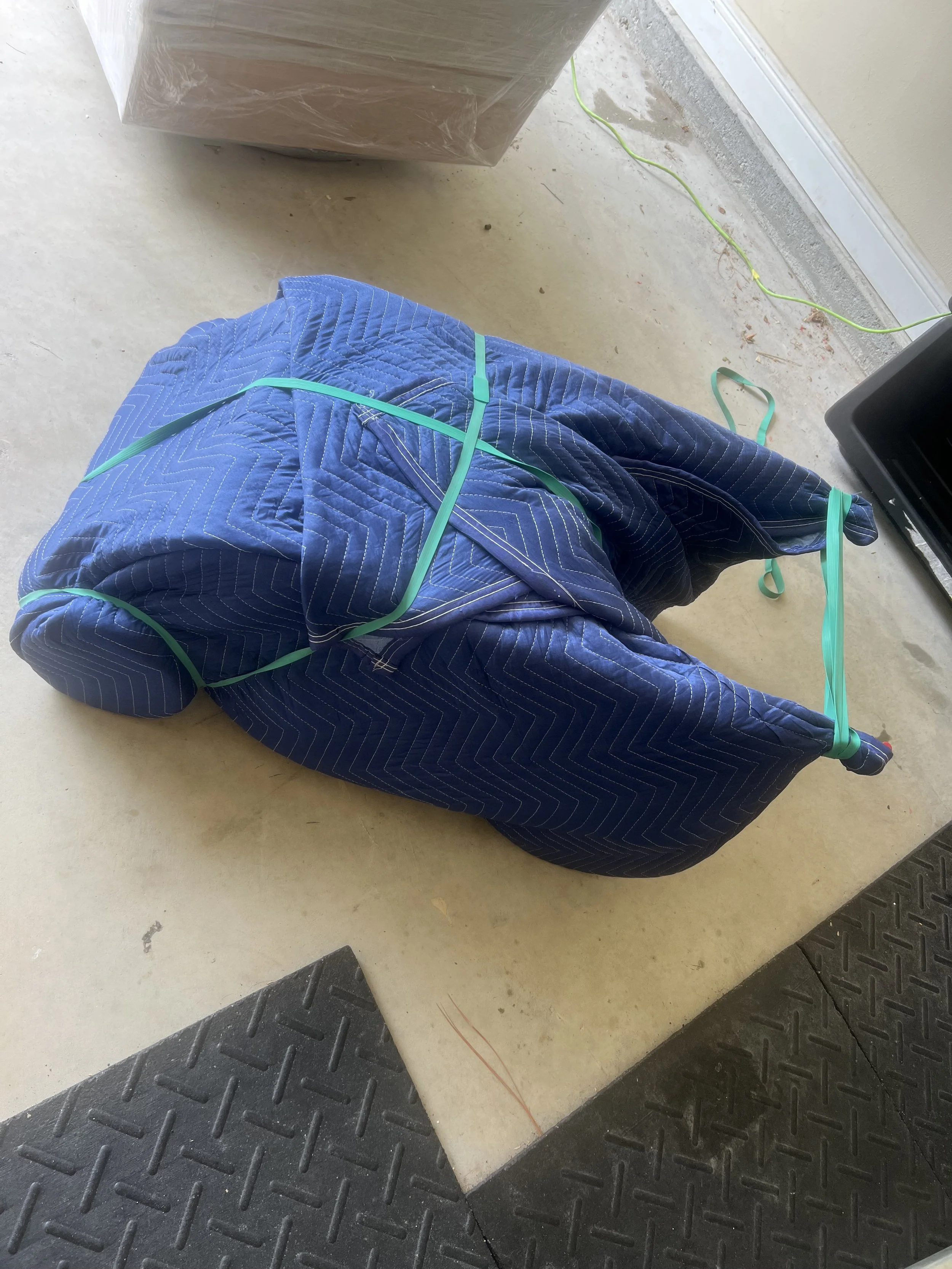 Floor with black rubber mats and a beige concrete surface. A large object wrapped in blue quilted moving blankets secured with green straps is in the foreground. In the background, there is a cardboard box, green extension cord, and a black object that could be a speaker or appliance.