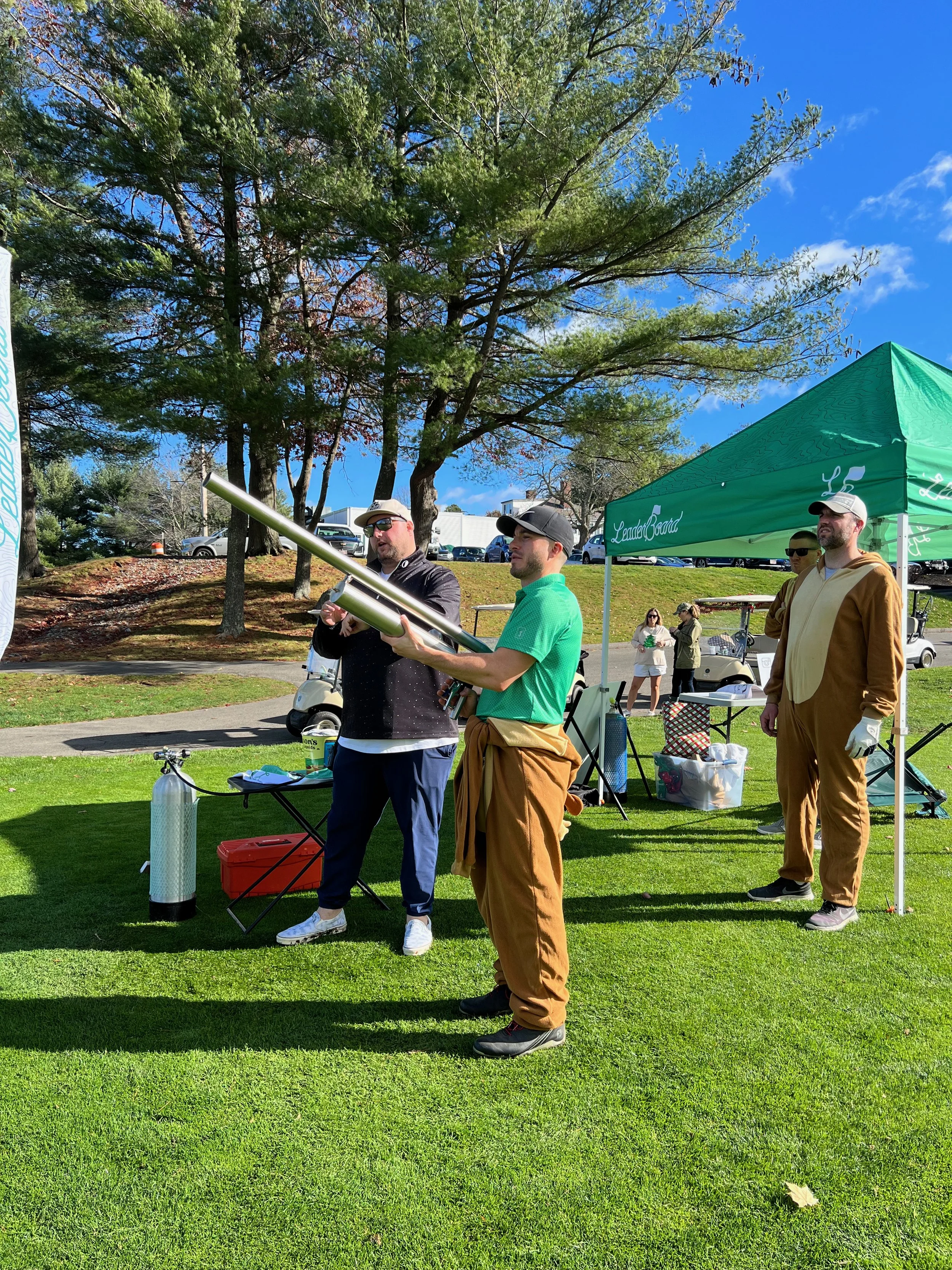 People gather on a sunny lawn beside a green Leander Board tent, golf carts, and event equipment.