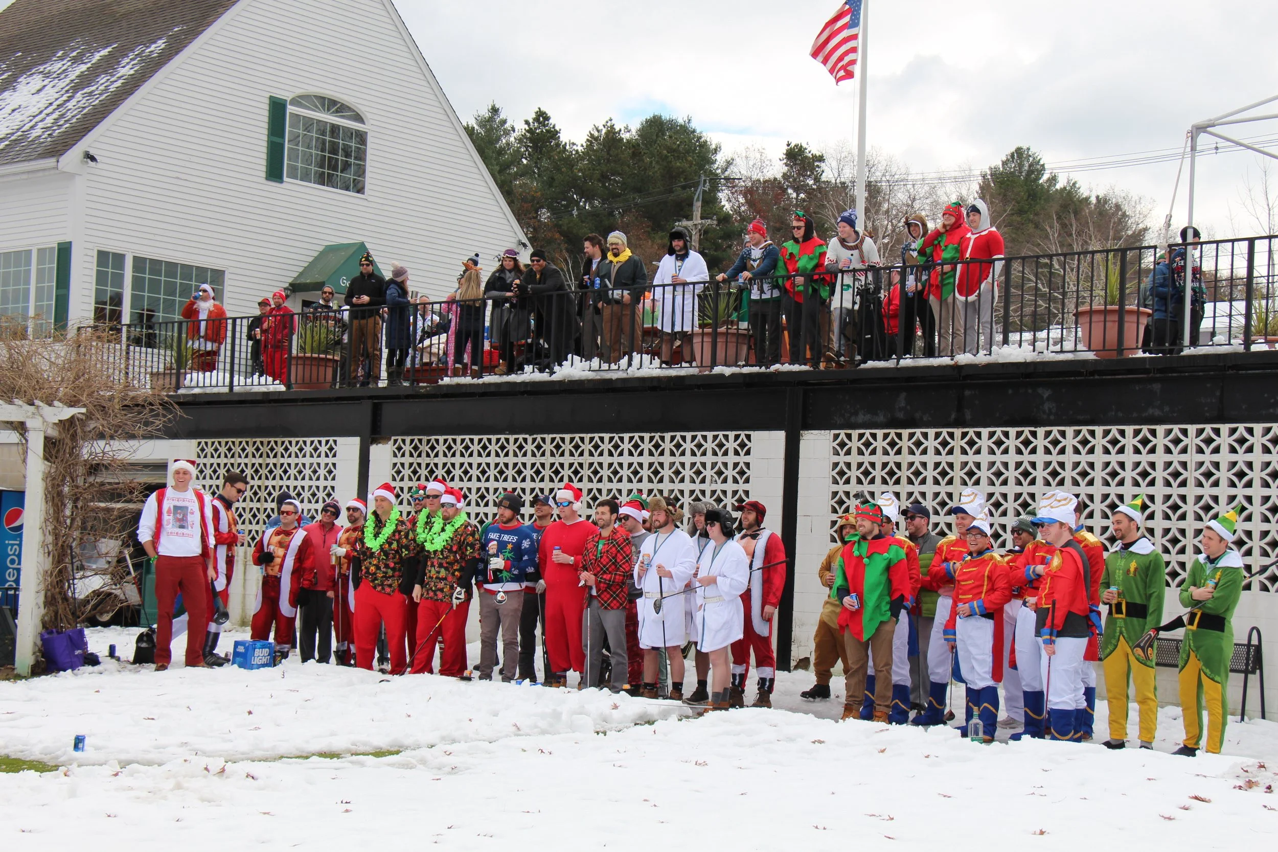 Large group in festive holiday costumes gathered in the snow outside a building with an American flag.