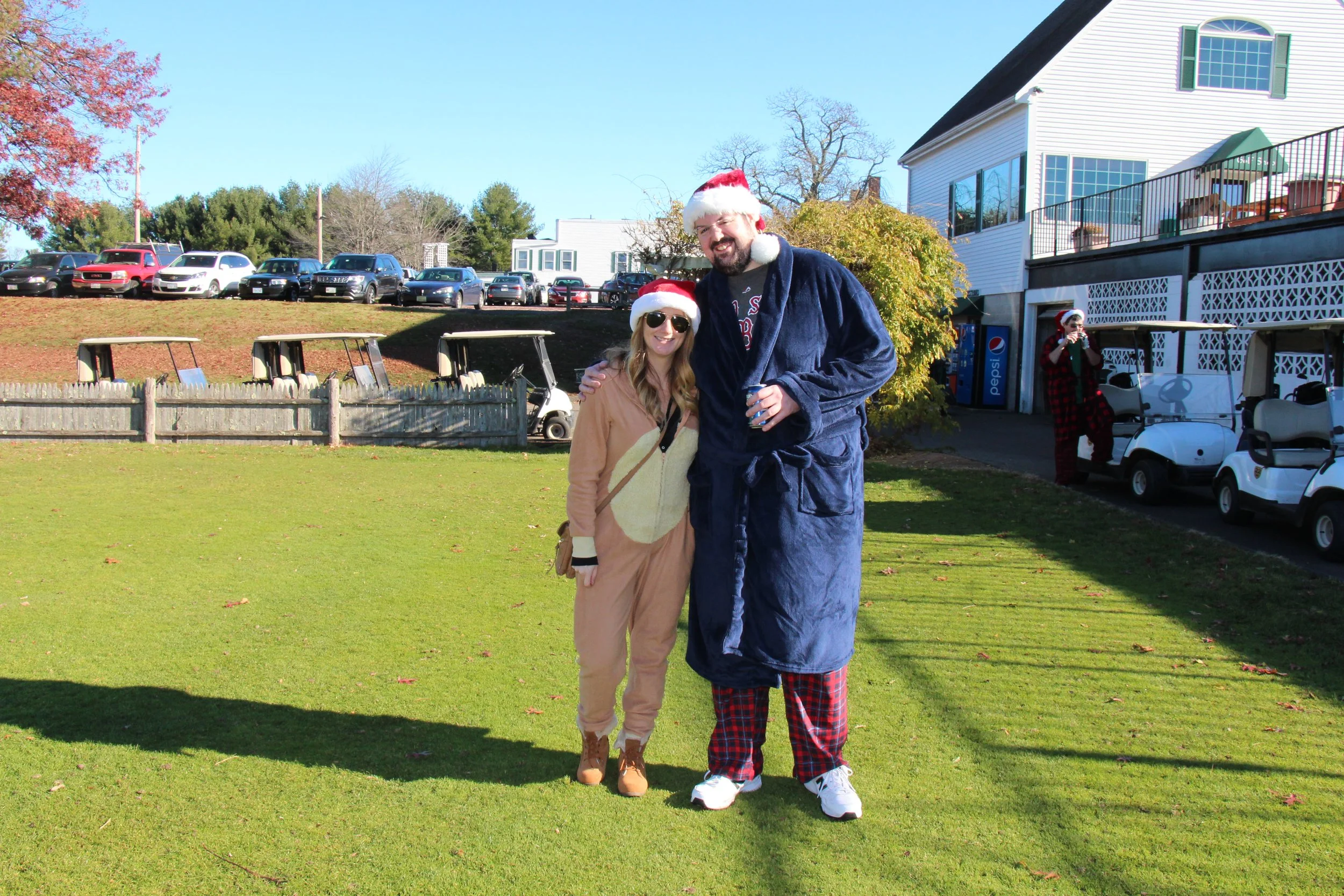 Two people wearing Santa hats pose on a sunny lawn beside golf carts and a house during a holiday gathering.