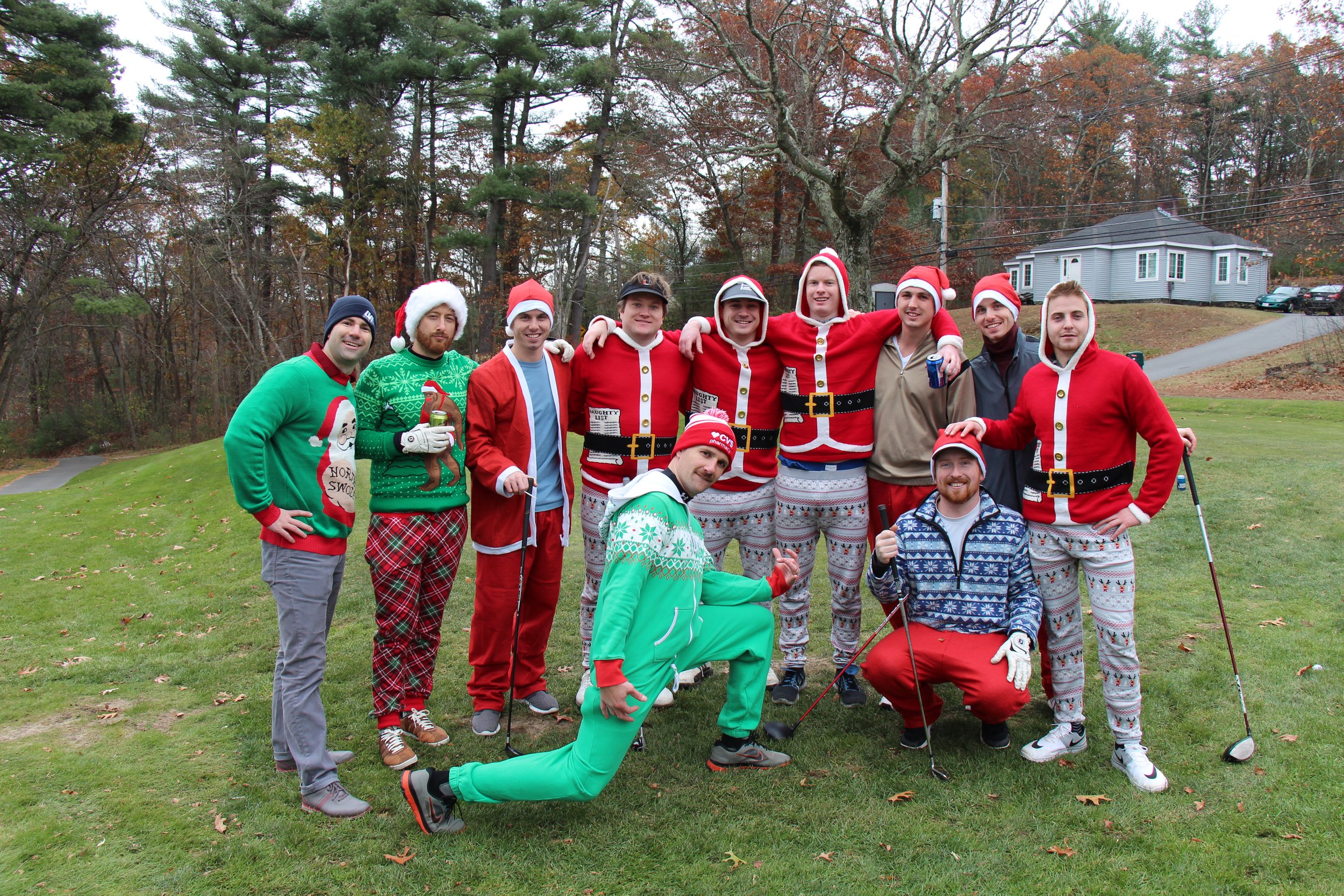 Group of friends wearing Santa costumes and festive Christmas sweaters posing outdoors on a lawn.