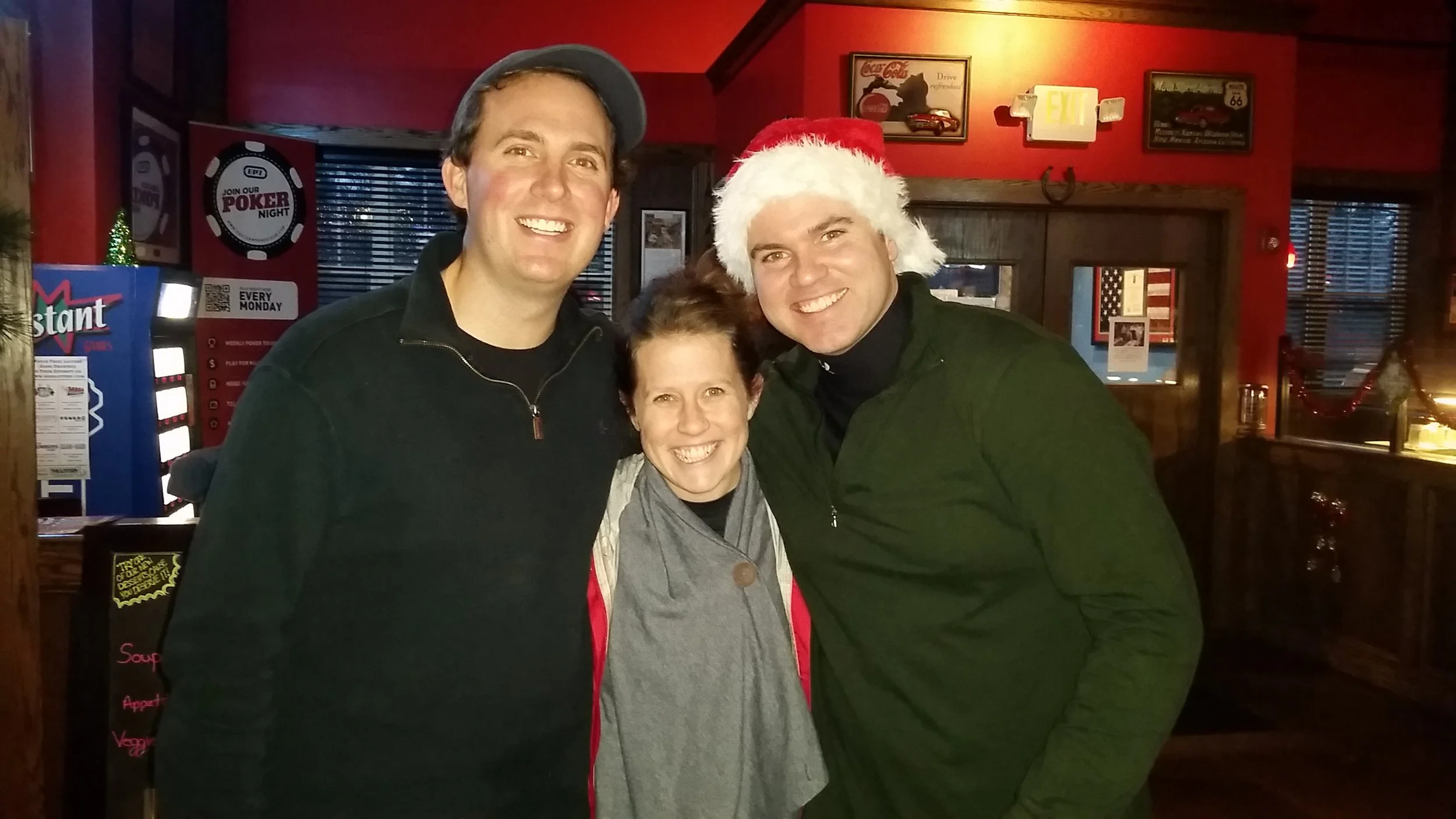 Three smiling people pose inside a warmly lit restaurant, with one person wearing a Santa hat.